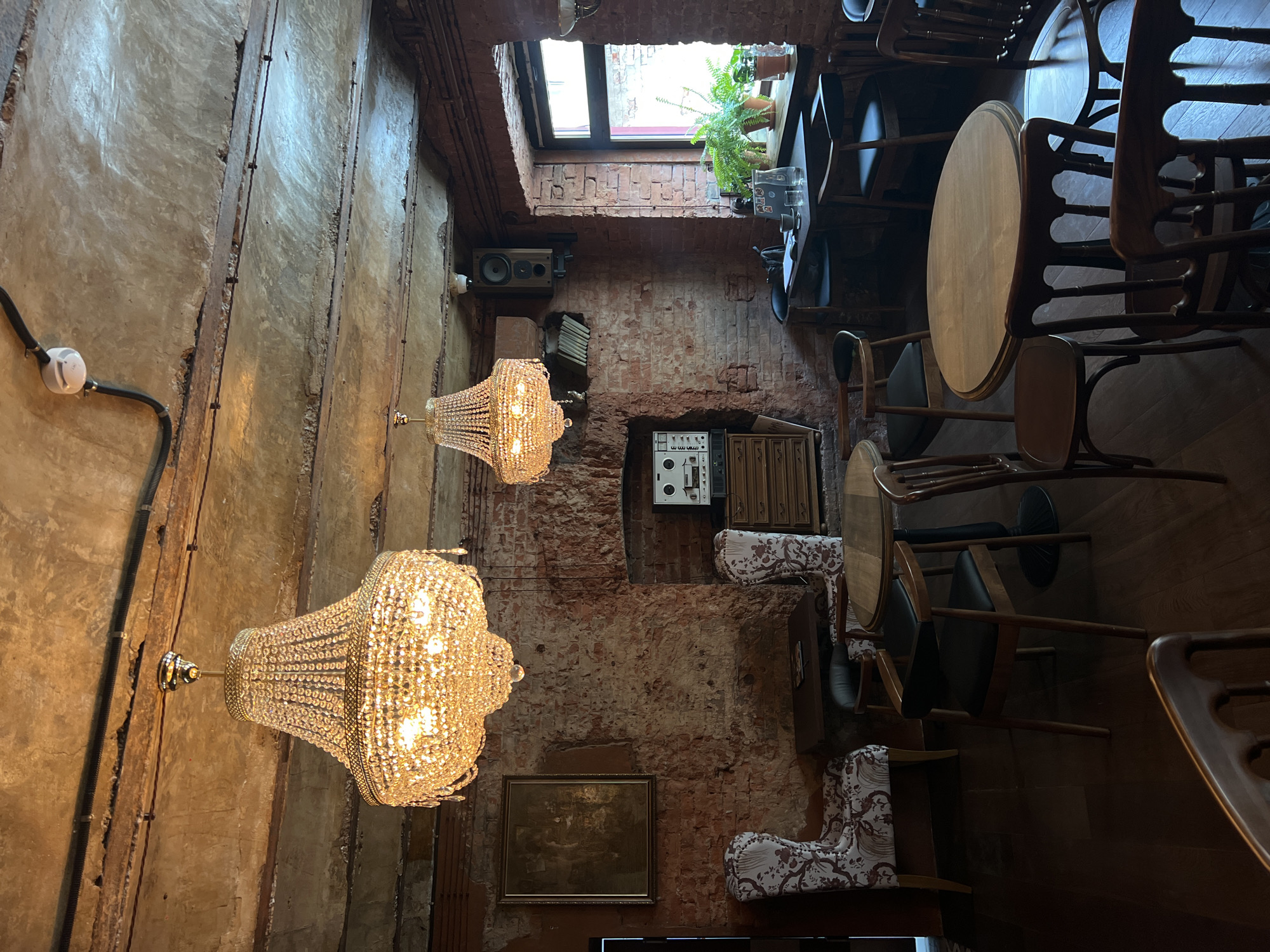 The image captures the interior of a building in Vladivostok, Russia. The walls are a mix of exposed brick and textured, aged concrete, with two ornate crystal chandeliers hanging from the ceiling. The ceiling also features a skylight framed by brickwork, revealing a glimpse of the outside and some potted plants. The floor is made of wood and is occupied by an assortment of wooden tables and chairs. A vintage sound system and equipment are mounted on the brick wall. Additionally, there is a framed painting. The building's interior design has a vintage aesthetic.