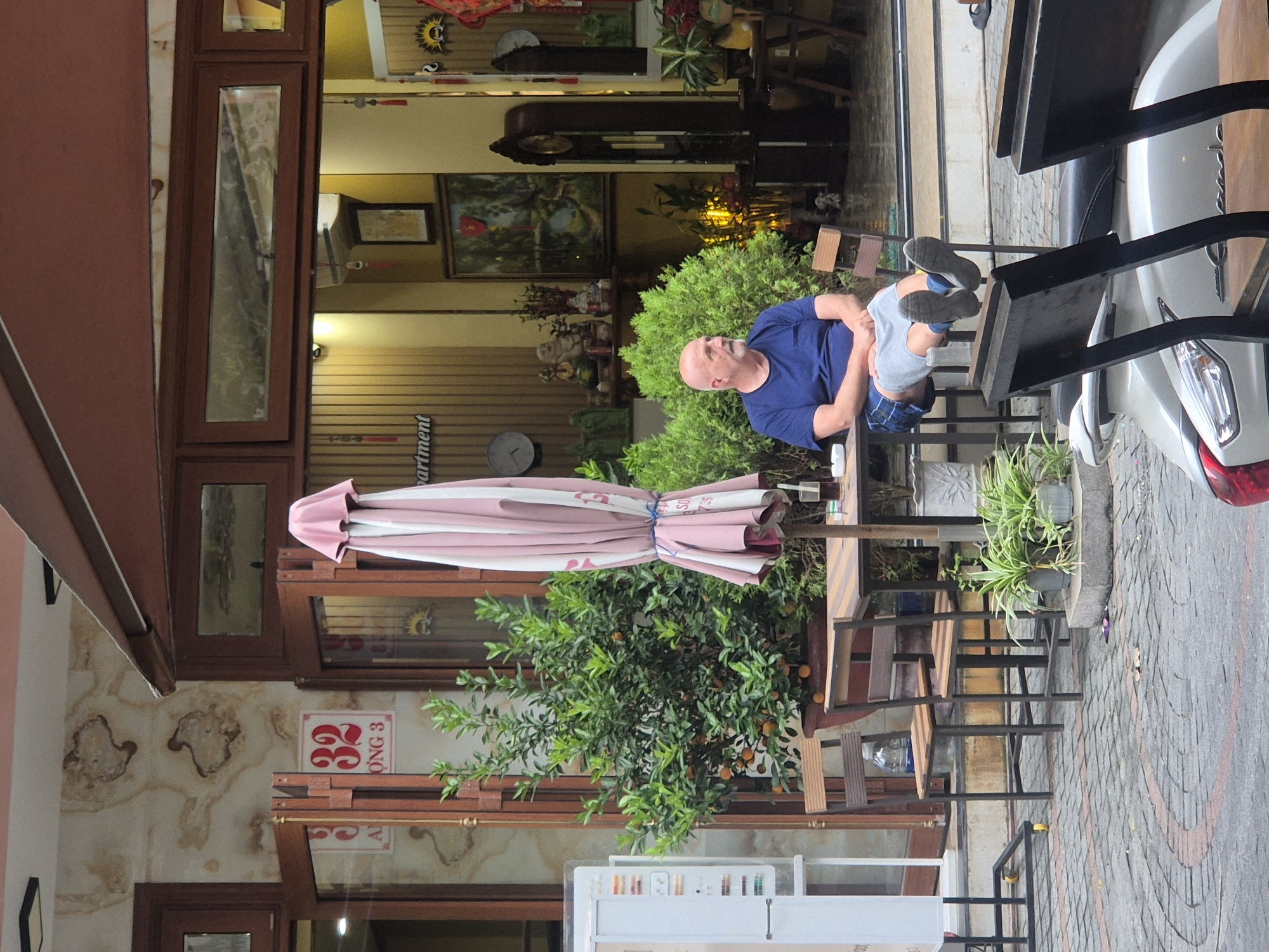 A bald, Caucasian man in a blue t-shirt and plaid shorts is seated at an outdoor cafe table. He is looking to his left, with his legs crossed and his feet resting on the rungs of his chair. A pink and white striped umbrella is open behind him, and a small orange tree is to his left. In the background, a street-level cafe has a warm yellow interior with several decorative items, including a grandfather clock and framed artwork. The cafe appears to be located on a cobblestone street in Đà Nẵng, Vietnam, indicated by the building number "32" and the Vietnamese characters on a sign.
