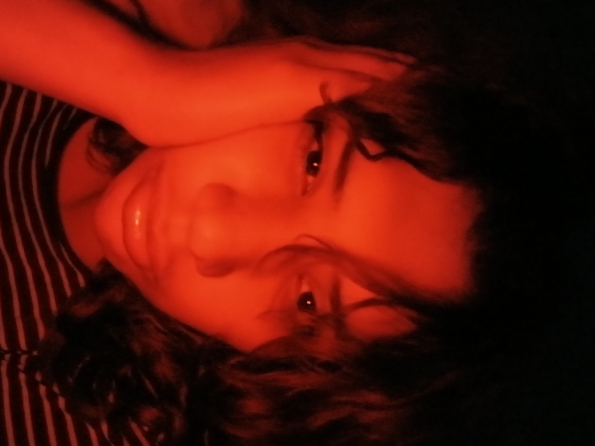 The image shows a person with dark curly hair, wearing a striped shirt. The person is holding their arm near their head. The lighting is predominantly red. The location is unknown but specified as Buenos Aires, Argentina. Without further information, a specific location in Buenos Aires cannot be determined.