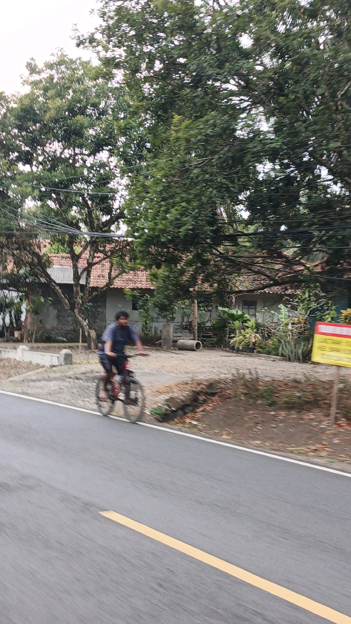 A young male is captured in motion, riding a bicycle on a paved road. He is wearing a dark blue t-shirt and dark shorts. The bicycle appears to be red. The road has a distinct yellow line marking its center. The background shows a rural or suburban setting with lush green trees and a house with a red tiled roof. A yellow sign with red markings is visible on the right side of the road. The image has a slightly blurred quality, suggesting movement. The scene is likely located in Cibenda, Indonesia, given the context provided.