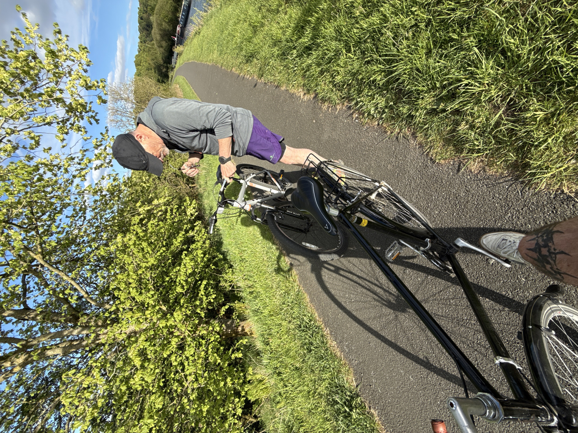 On a sunny day, a man in a gray hoodie, purple shorts, and a black baseball cap is bending over, appearing to adjust something on a bicycle. The bicycle is lying on its side on a paved path. The path is bordered by green grass on both sides, with trees and foliage visible in the background and to the side. The man is focused on the bicycle, and the setting is outdoors, possibly along a canal based on the canal boats visible in the background of the image. Part of another bicycle and a person's leg with a tattoo is visible in the foreground.