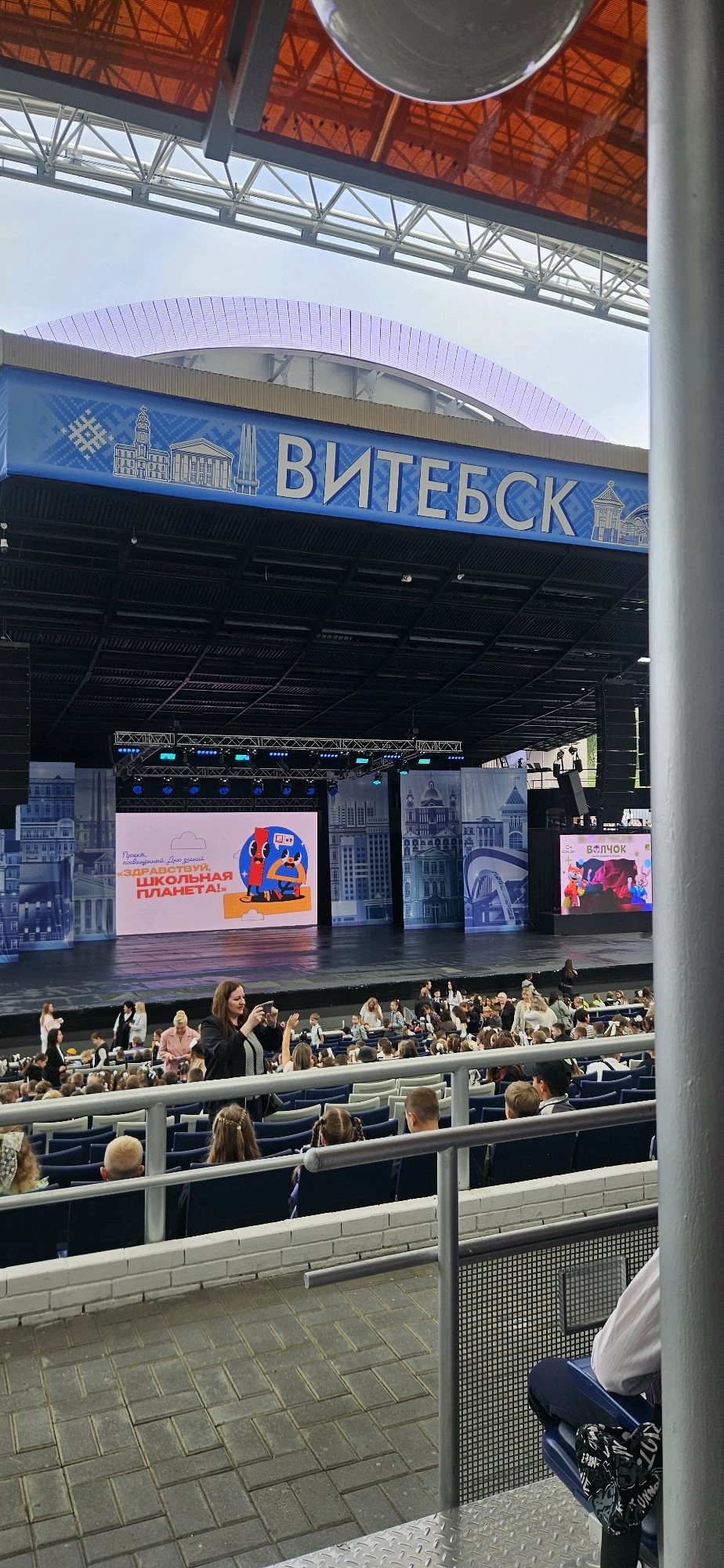 An outdoor stage is set up for a performance. The stage has a large screen displaying a colorful graphic and text that reads "ЗДРАВСТВУЙ, ШКОЛЬНАЯ ПЛАНЕТА!". The name of the city, "ВИТЕБСК", is prominently displayed in large blue letters on a blue banner above the stage. The banner also features white decorative elements and images of buildings. The stage is lit by stage lights, and speakers are visible on either side. In front of the stage, an audience, mostly composed of children, is seated in rows of blue seats. Some adults are also present, including a woman in the foreground holding a camera. The seating area is paved with gray interlocking stones. The overall scene suggests an event is taking place at an open-air venue in Vitsebsk, Belarus.