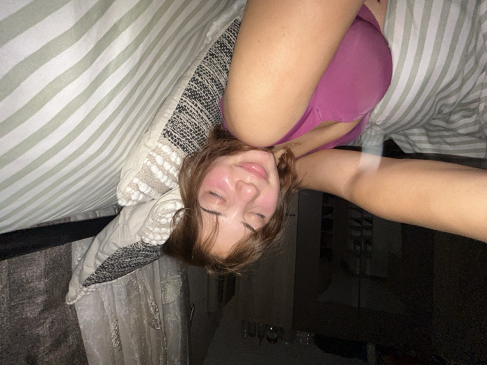 A young woman lies upside down in bed, eyes closed, wearing a pink bra. Pillows with varying textures and patterns surround her head. A striped duvet covers a portion of the scene. Part of an arm is visible extended to the side. Behind her, a dark area suggests a room or hallway, with glimpses of a window or reflective surface. The overall impression is casual and relaxed.