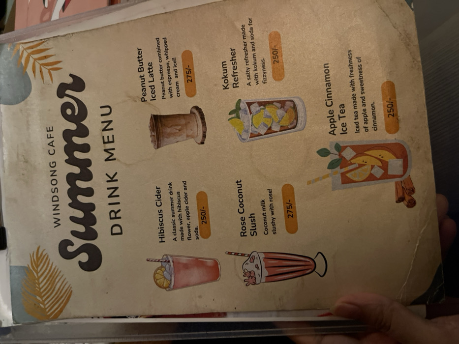 The image displays a close-up view of a laminated "Summer DRINK MENU" from "WINDSONG CAFE" in Dibrugarh, India, held by a person's hand. The menu features a light brown, aged paper background with illustrations and text.
The menu lists five distinct summer beverages, each with a description and price:
1. **Peanut Butter Iced Latte:** Described as "Peanut butter combined with espresso, whipped cream and ice!!" priced at 275/-. An illustration of a tall, light brown drink with a frothy top in a wooden-textured glass accompanies it.
2. **Kokum Refresher:** A "salty refresher made with kokum and soda for fizzyness," available for 250/-. This is depicted as an iced drink with lime slices in a clear glass.
3. **Apple Cinnamon Ice Tea:** An "Iced tea made with freshness of apple and sweetness of cinnamon," priced at 250/-. The illustration shows a glass of iced tea with apple slices, mint, and cinnamon sticks.
4. **Hibiscus Cider:** A "classic summer drink made with hibiscus flower, apple cider and soda," costing 250/-. It's shown as a red iced drink with a lemon slice and straw in a tall glass.
5. **Rose Coconut Slush:** "Coconut milk slushy with rose!" priced at 275/-. An illustration of a layered pink and white slushy with whipped cream and sprinkles in a fluted glass accompanies it.
The menu is designed with a summer theme, indicated by the large "Summer" title and subtle leaf motifs on the background. The overall impression is a vibrant selection of refreshing drinks offered by the cafe.