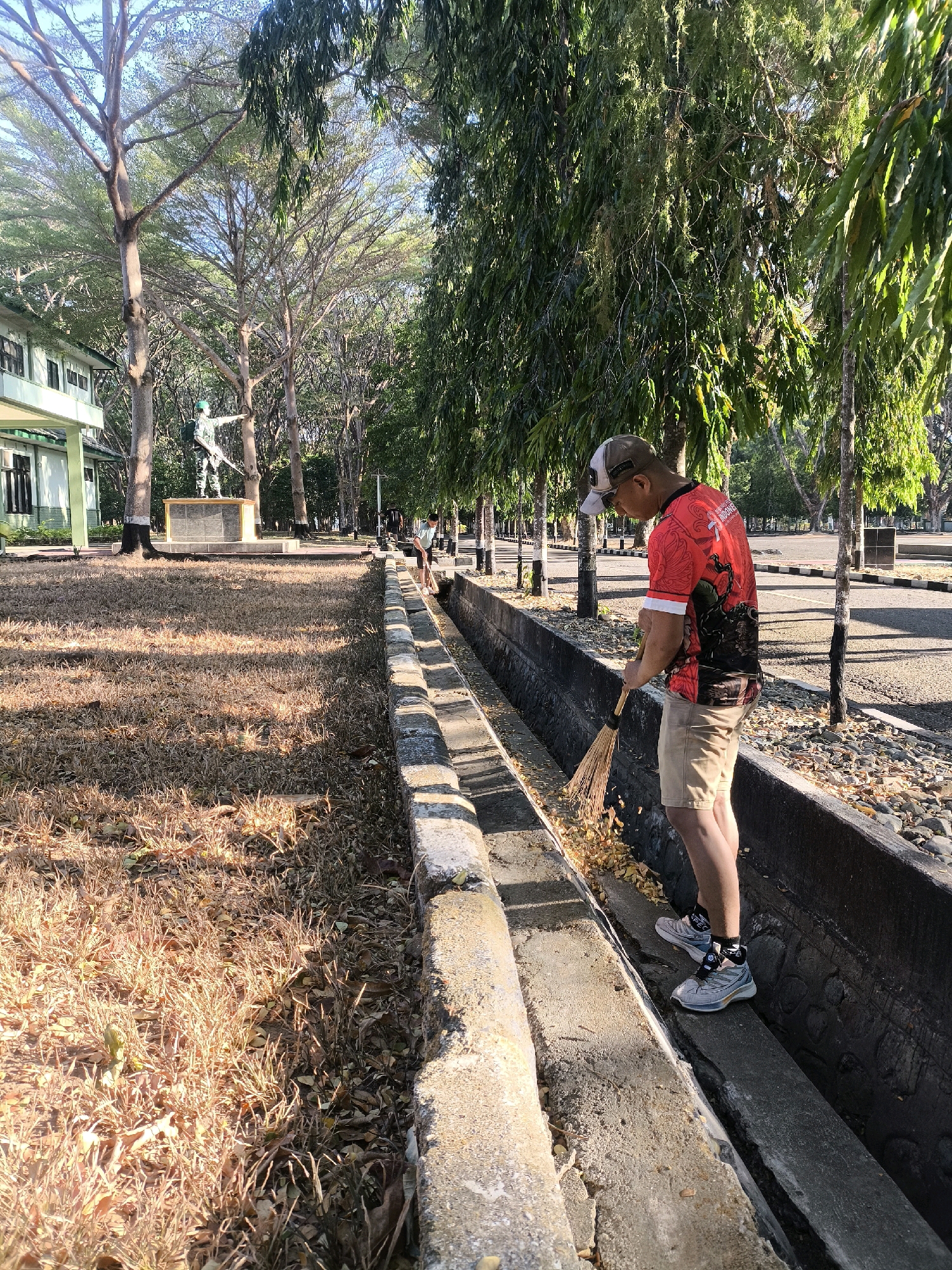 A man is sweeping leaves in a concrete ditch. He is wearing a red shirt, khaki shorts, and a baseball cap. The ditch is lined with concrete, and there are trees and a building in the background. The ground is covered in dry grass and leaves.