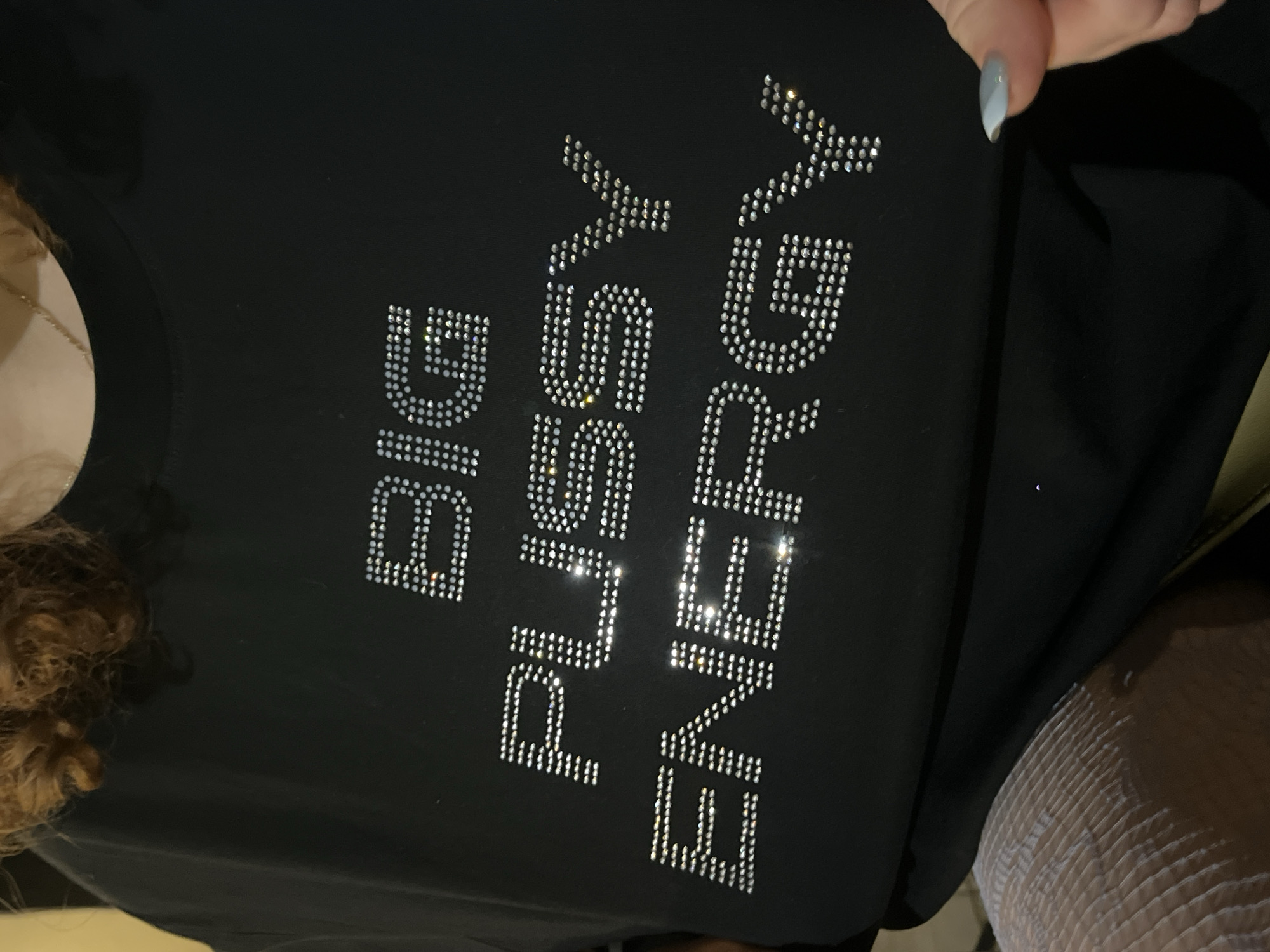 A close-up shot focuses on a black garment adorned with the phrase "BIG PUSSY ENERGY" in sparkling rhinestones. The text is arranged vertically, with "BIG" at the top, followed by "PUSSY," and then "ENERGY." To the left, a glimpse of curly hair and a gold chain is visible. A hand with a light blue and white nail is near the top right corner. A texture resembling a white, net-like material is at the bottom right of the image. The scene does not appear to be located in Tel-Aviv, Israel, as there are no defining characteristics of that region in the image.