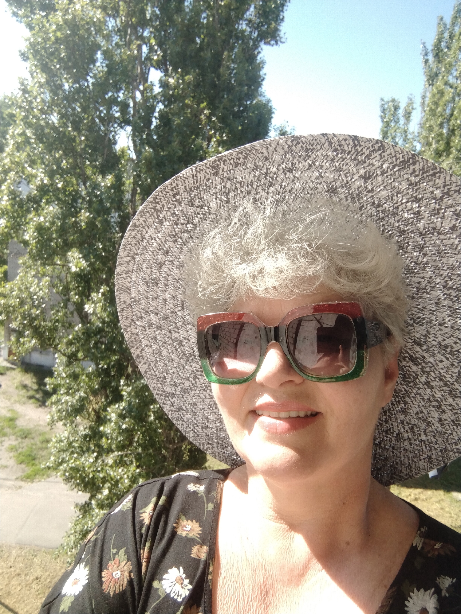 A woman is shown in a close-up selfie, wearing a wide-brimmed hat and sunglasses. She is wearing a dark floral-patterned top. Behind her is a background of green trees, suggesting an outdoor setting. Based on the visual content, determining the precise location in Horishni Plavni, Ukraine, is impossible.
