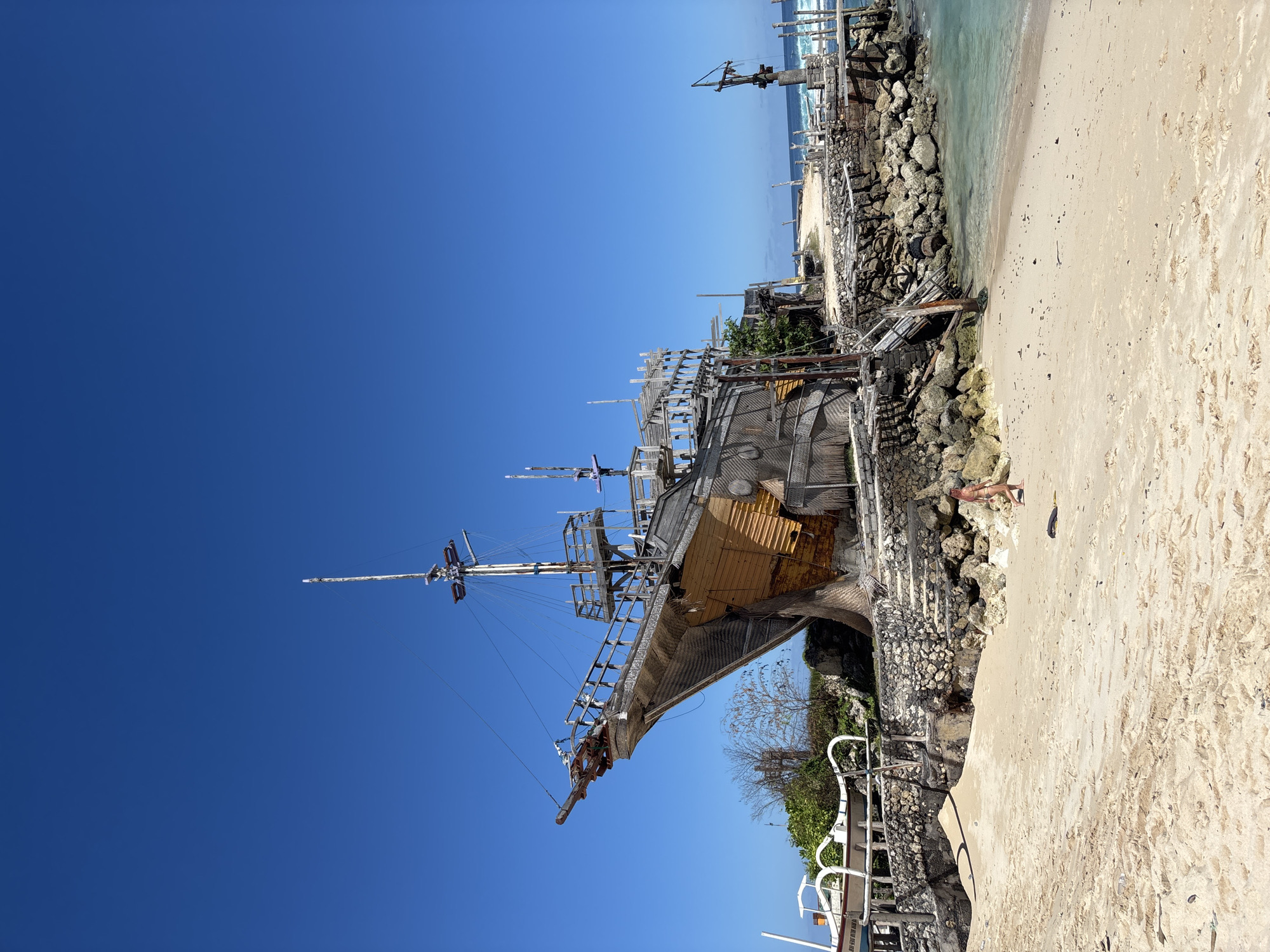 The image shows a beached wooden ship with multiple masts in the city of Kuta Selatan, Indonesia. The ship sits next to a sandy beach with a rocky shoreline. A person is walking along the beach. The sky is a clear, bright blue. The ship is positioned to the left of the frame. The ocean is visible in the background. A white railing runs along the bottom right of the photo. Vegetation can be seen near the ship.