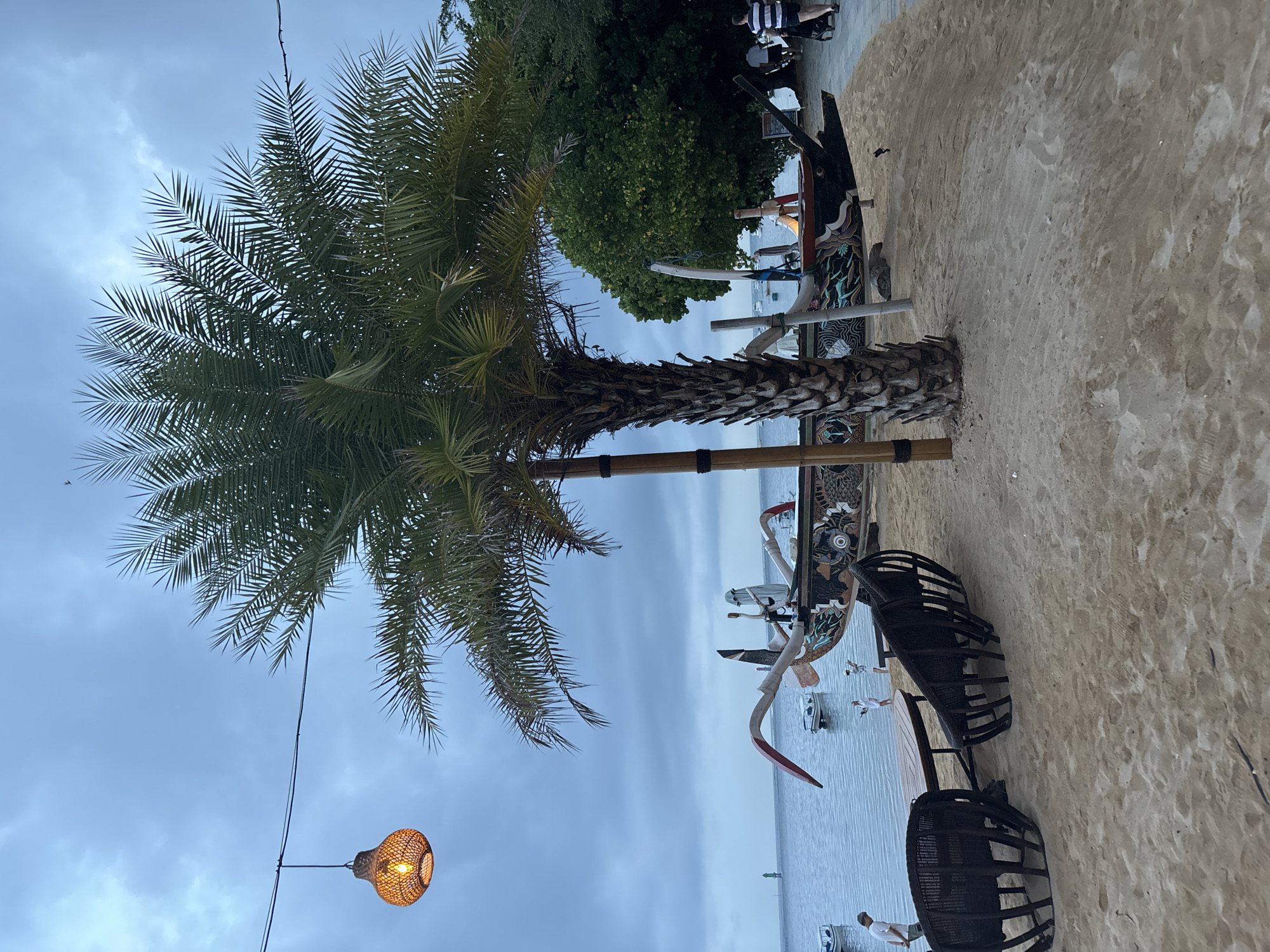 The image depicts a beachfront scene in Denpasar, Indonesia, with a focus on a sandy area leading to the ocean. In the foreground, a round wooden table is flanked by two dark, open-weave chairs made of rattan or similar material, positioned on the sand. Behind this seating arrangement, a prominently featured palm tree stands tall, its fronds extending towards the overcast sky.
A traditional Indonesian fishing boat, known as a jukung, is displayed on the sand, positioned diagonally across the frame. This boat is elaborately decorated with colorful carvings and motifs. Several smaller boats are visible on the calm blue water of the ocean in the background, along with a green channel marker.
To the left of the scene, a man in a white shirt and tan shorts stands near the water's edge. To the right, a couple walks along a paved pathway near some trees. A decorative woven pendant light hangs from a wire above the scene, casting a warm glow. The overall atmosphere suggests a relaxed, coastal setting, possibly a restaurant or tourist area.