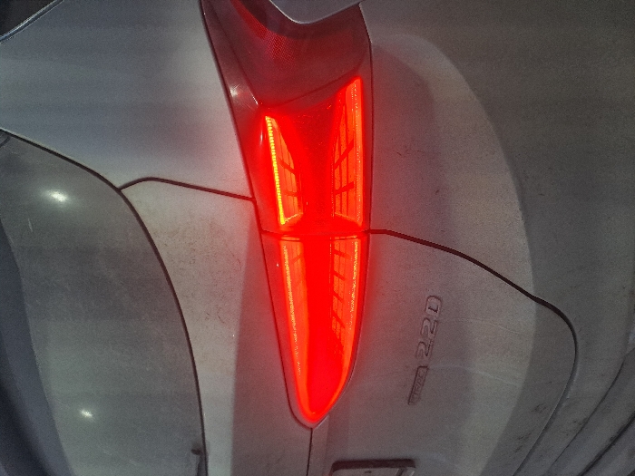 A close-up, slightly blurry, angled shot shows the rear taillight of a dusty gray vehicle. The taillight is bright red and vertically oriented, with a distinct division in the middle. "CRDi 2.2D" is visible in raised lettering on the car's body near the light. The vehicle's panels are visibly dirty, with some marks and smudges across the surface.