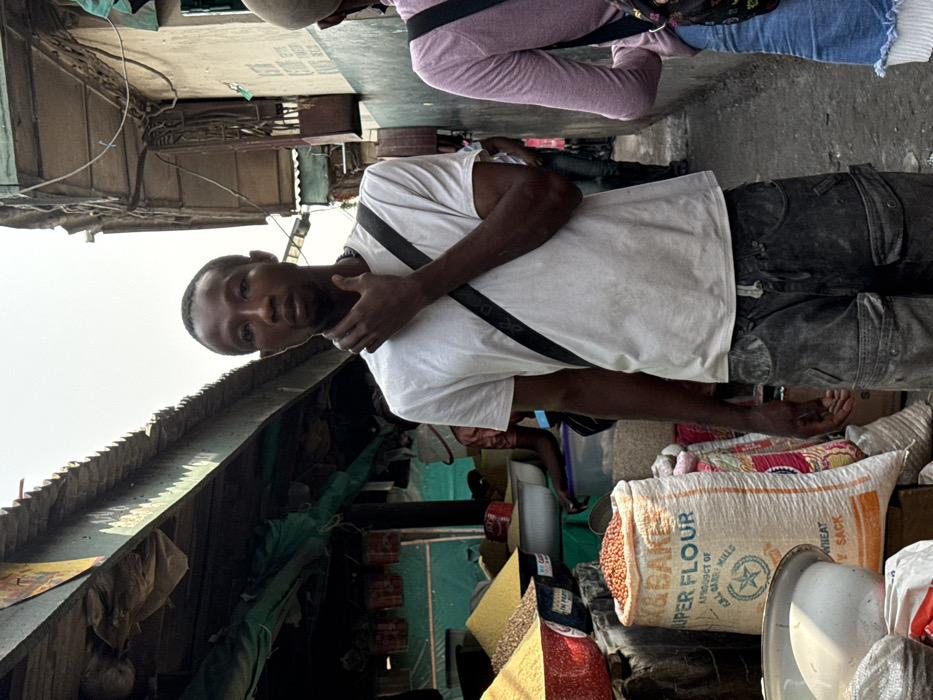 This image depicts a narrow, bustling outdoor market alleyway in Lekki, Nigeria. A dark-skinned male, appearing to be in his 20s or 30s, stands centrally, facing the camera with a neutral expression. He wears a white short-sleeved t-shirt, dark, worn cargo pants, and a black cross-body bag across his chest. His right hand is raised, with fingers curled, touching his chin and jawline. To his immediate left, market stalls are set up under a green fabric awning and corrugated metal roof. These stalls display various bulk dry goods in open containers and sacks, including a large white sack labeled "D&BAKER SUPER FLOUR" filled with orange-brown beans or legumes. Other containers hold yellow, white, and brown granular products. To the right, another individual is partially visible from the back, wearing a light pink long-sleeved top, blue denim shorts with a fringed hem, and a patterned backpack, walking away from the camera. The right side of the alley is bordered by an aged, light-colored building wall featuring exposed electrical conduits and wiring. The ground is a worn, uneven surface. The scene is illuminated by diffused daylight.