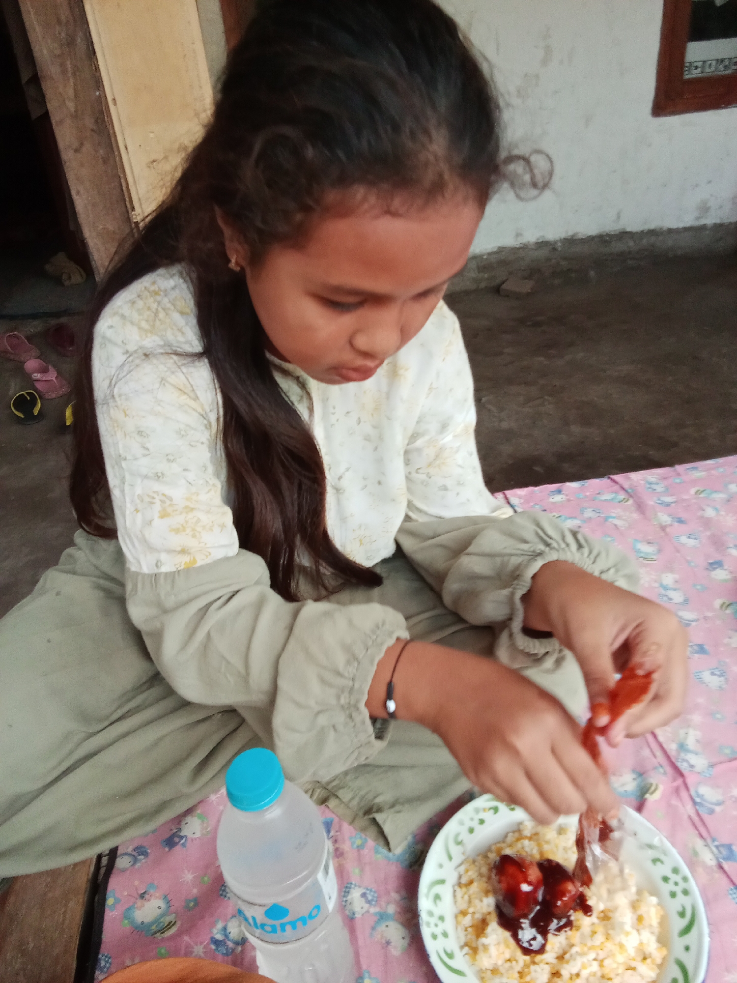 A young girl with long, dark hair is sitting on the ground in what appears to be an outdoor area, possibly a yard or veranda, in Selowogo, Indonesia. She is wearing a long-sleeved, light-colored shirt with a floral pattern and light olive green pants. Her head is bowed as she focuses on a bowl of food in front of her. The bowl contains what looks like scrambled eggs topped with dark red, possibly meat, items coated in sauce. In her hands, she is holding a piece of food, appearing to be adding it to the bowl. To her left, a plastic bottle of water with a blue cap is visible. The ground is dirt and concrete. In the background, there is a wooden structure and a wall. There are also a few pairs of children's sandals on the ground to the left of the girl. The overall scene suggests a casual, informal mealtime.