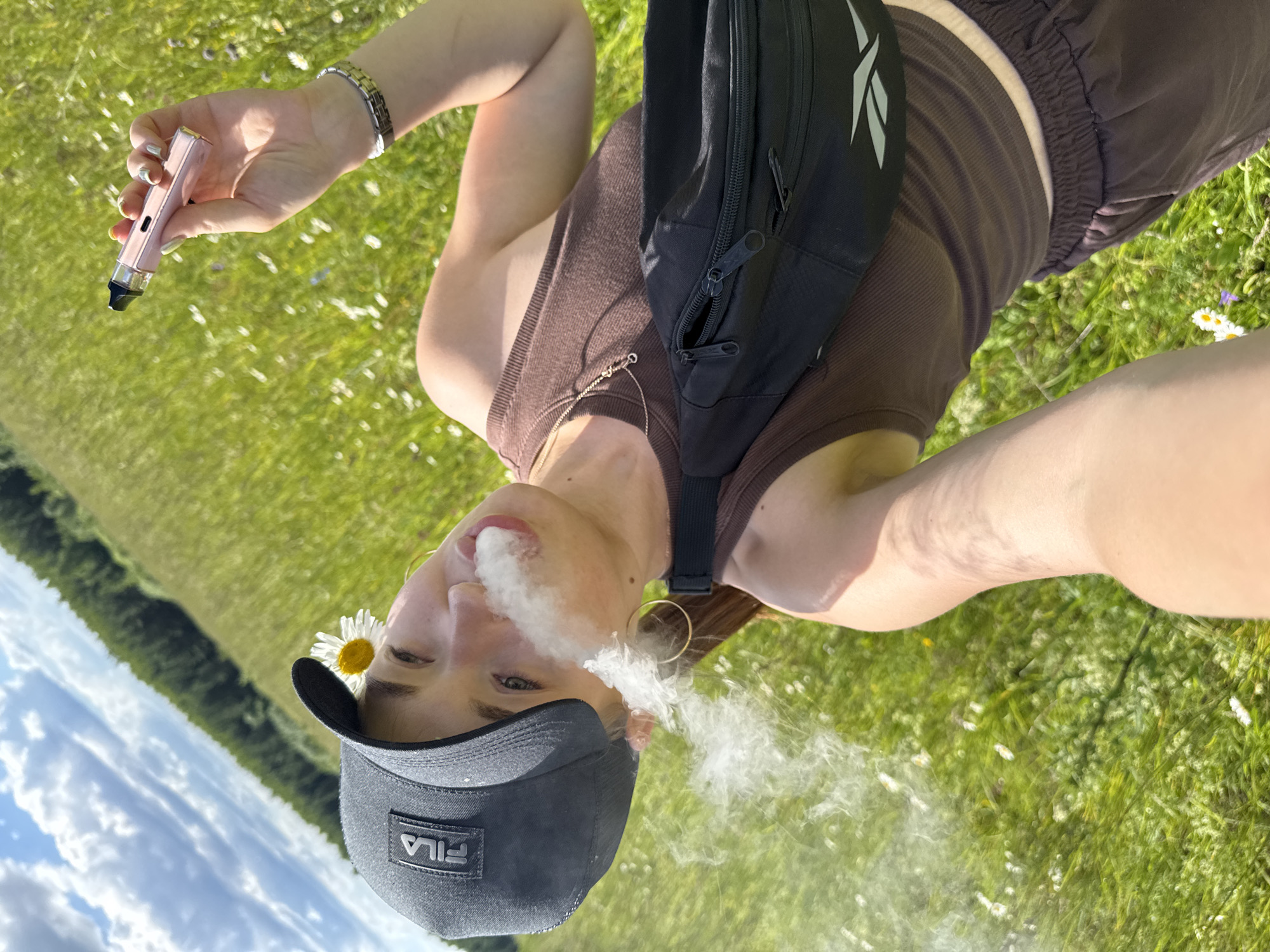 A person is taking a selfie in a grassy field with trees in the background under a cloudy sky. The person is wearing a black Fila cap, a brown tank top, brown sweatpants, and a black fanny pack. They are holding a pink vaping device and exhaling a cloud of smoke. A daisy is tucked behind the person's ear. The location is identified as the village of Garevaya, Russia.