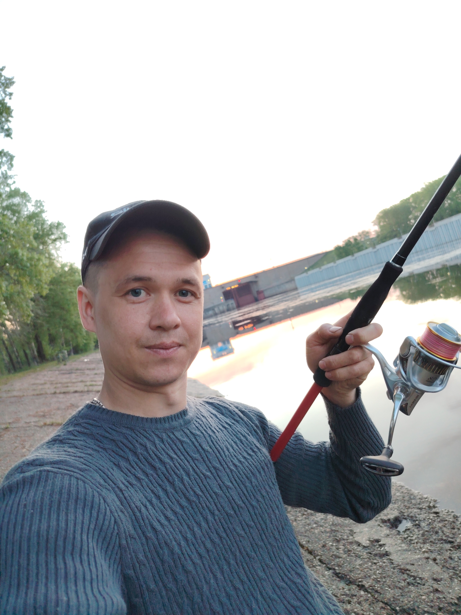 A man is holding a fishing rod next to a body of water. The man is wearing a gray sweater and a black cap. The fishing rod is black with a silver reel. The water is calm and reflects the buildings in the background. The location is the city of Chaikovsky, Russia.