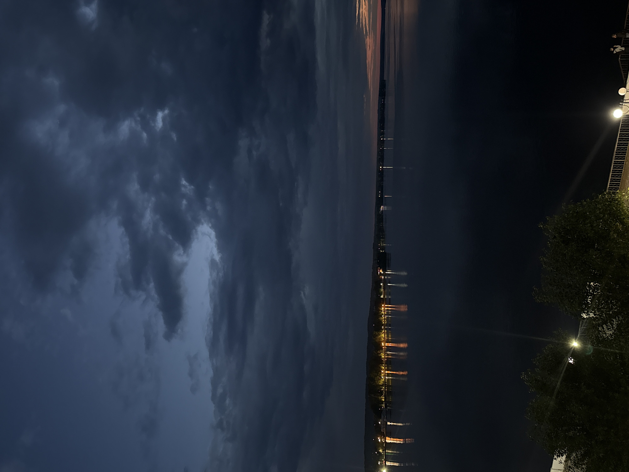 The image shows a twilight view of the Kama River in Votkinsk, Russia. The sky is filled with dark, heavy clouds, suggesting a storm or the end of a cloudy day. The river is dark and still, reflecting the lights from the shore. On the far bank, a line of lights runs along the horizon, indicating buildings or a town. In the foreground, there are trees and a railing, suggesting that the photo was taken from a balcony or elevated walkway. Two figures, a man and a woman, are standing near the railing, looking out at the river. The overall atmosphere is moody and serene, with the interplay of dark clouds, calm water, and distant lights.
