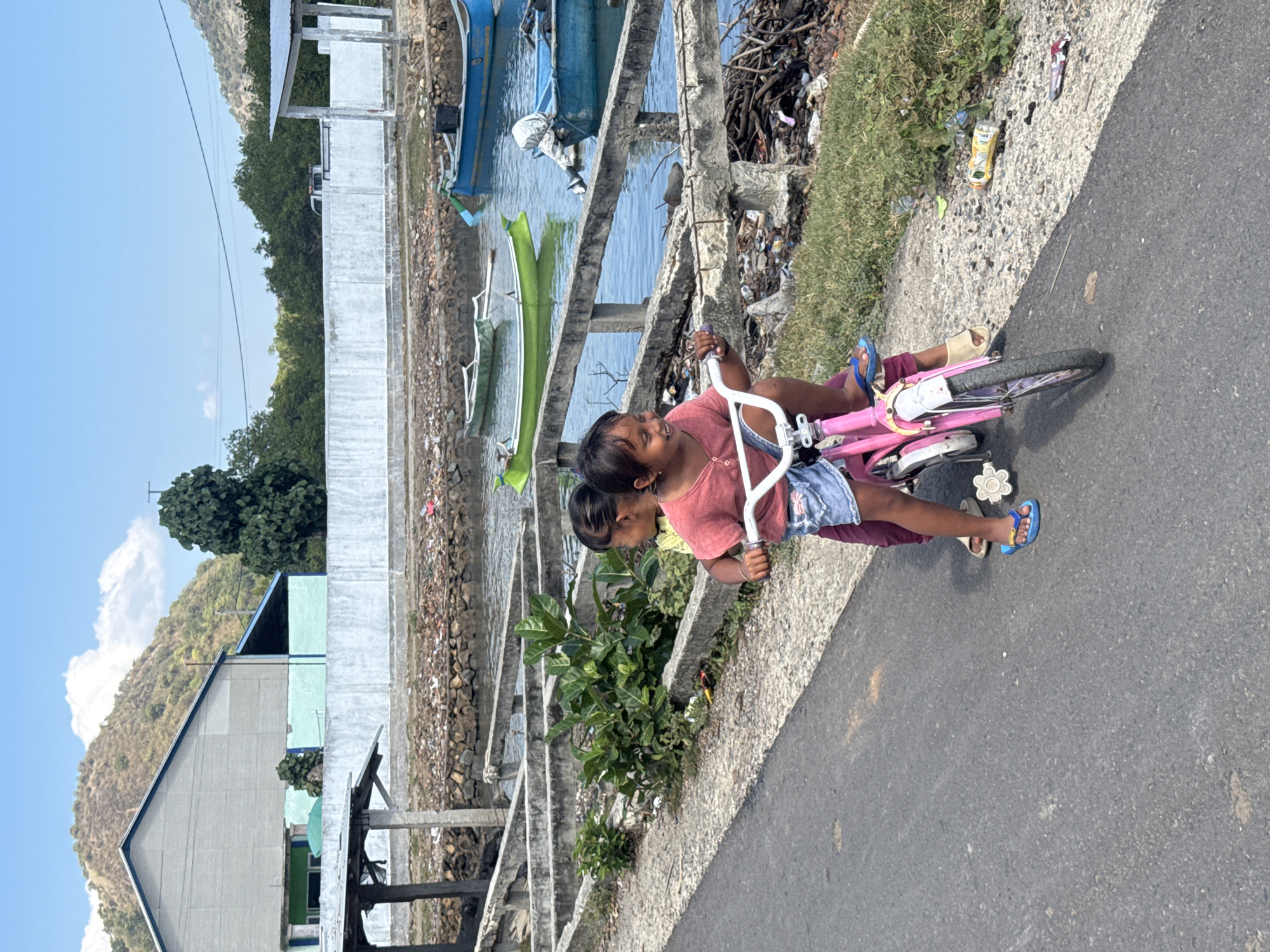 In Kertasari, Indonesia, two young girls ride a pink bicycle on an asphalt road. The girl in front wears a red t-shirt and denim shorts, while the girl behind her is partially visible, wearing a yellow shirt. They are near a body of water, possibly a harbor, where several boats are moored. The background features a textured wall, lush green vegetation, and rolling hills under a clear blue sky with scattered clouds. Debris is visible along the waterline and on the grassy embankment.