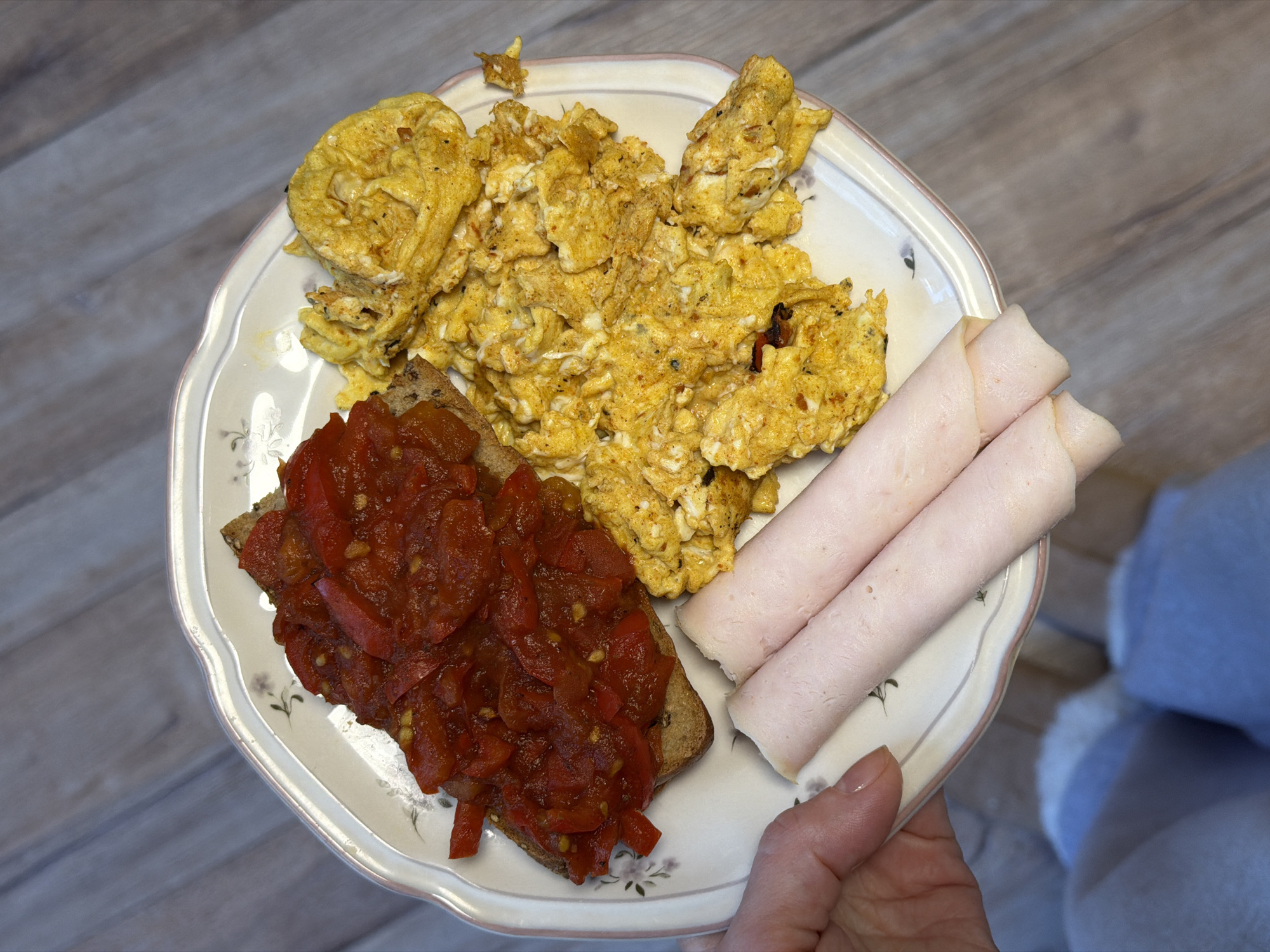 A plate of food is held in someone's hand, likely indoors. The plate contains scrambled eggs, a slice of bread topped with a red vegetable mixture, and two rolled slices of ham. The background shows a light-colored surface, and part of a blue fabric (likely clothing) is visible. There are no visible indications that the location is Perm, Russia.