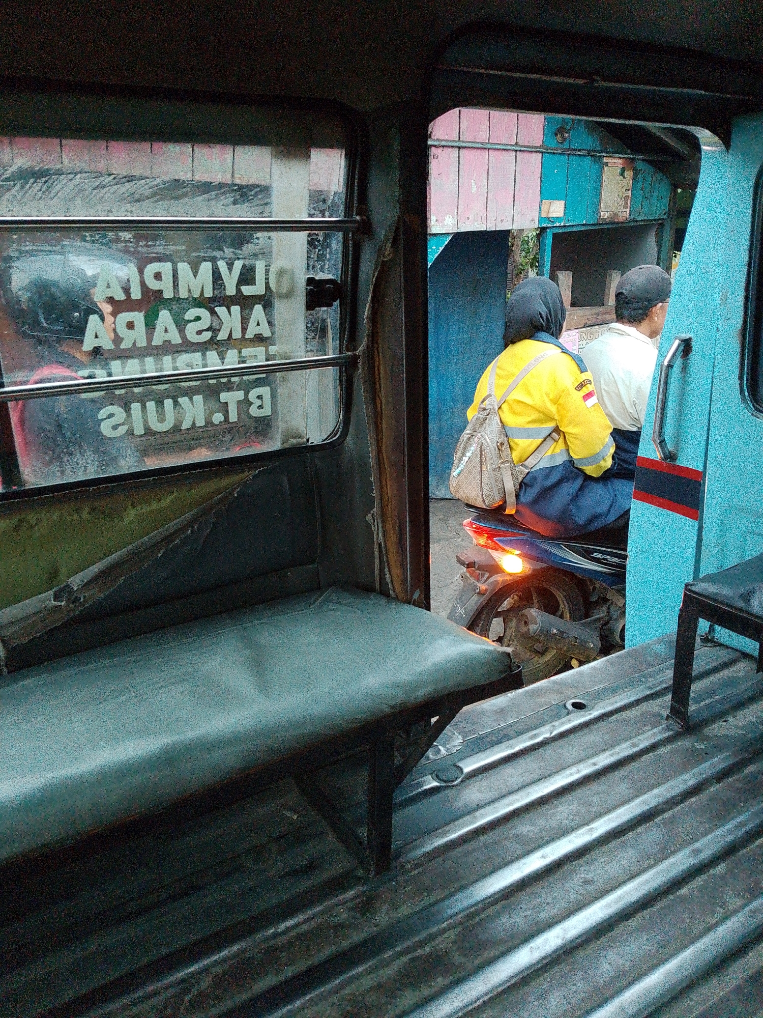 The image shows the interior of a vehicle, likely a bemo or a similar public transport vehicle, in Bandar Khalipah, Indonesia. Through the open side door, two passengers are visible on a motorcycle. The passenger in front is a woman wearing a yellow and blue uniform, with a backpack slung over her shoulder. She is wearing a black hijab. Behind her, a man wearing a black cap is seated. They are stationary, with the motorcycle's brake light illuminated.
Inside the vehicle, a worn green bench seat is visible in the foreground. The interior walls are covered with a reflective material, and metal bars run across the windows. Text on the window is partially visible and appears to be reversed, suggesting it is seen through reflection. The exterior environment shows building structures with pink and blue walls. The floor of the vehicle is made of corrugated metal. The overall impression is one of a typical scene in a busy urban environment in Indonesia.