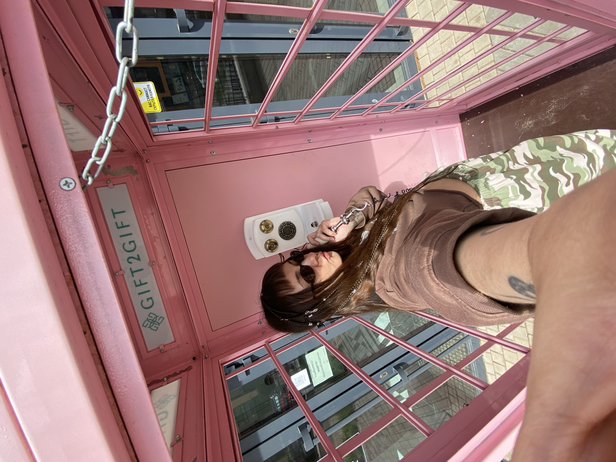 A woman is taking a selfie inside a pink phone booth, holding a landline phone handset to her ear. She is wearing sunglasses, a brown sweatshirt, and camouflage-patterned pants. The phone booth is decorated with a sign that says "GIFT2GIFT" and has a chain attached to the top. A sign on the outside of the booth reads "ВАС СНИМАЕТ СКРЫТАЯ КАМЕРА" (You are being filmed by a hidden camera). Another sign inside the booth says "Улыбнитесь ведется видеонаблюдение :)" (Smile, video surveillance is in progress :)). The phone booth is located outside a building with glass windows, and a patterned sidewalk is visible. The image was taken in Izhevsk, Russia.
