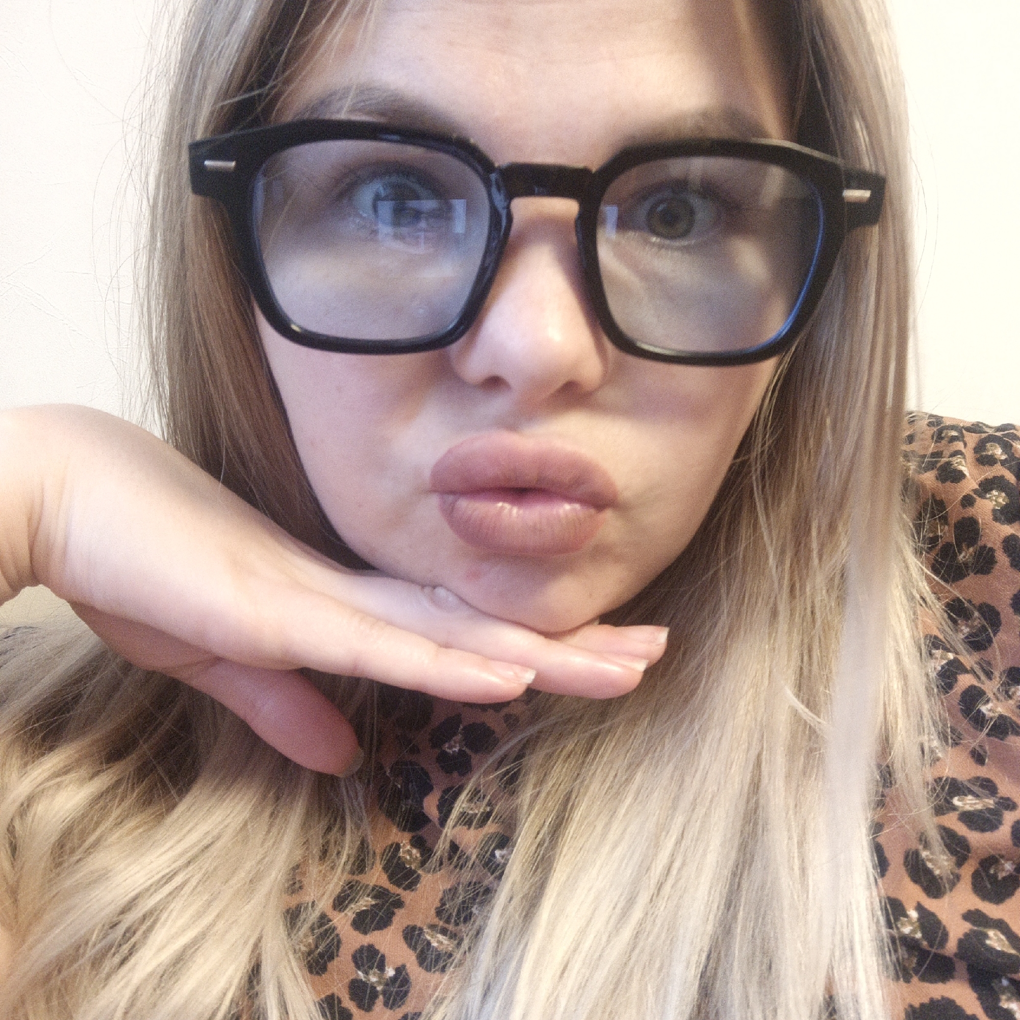 A close-up selfie shows a woman from the chest up, centered in the frame. She has long, blonde hair, possibly with darker roots, that falls around her face and over her shoulders. She wears prominent black, thick-framed rectangular eyeglasses, through which her eyes appear wide and slightly surprised, with reflections visible on the lenses.
Her chin rests on her right hand, which is cupped under her jawline. Her lips are pursed into a distinct "duck face" or pout, enhanced with a pinkish-nude lipstick. The skin on her face and hand is fair, with natural-looking fingernails.
She is dressed in a top featuring a brown and black leopard print pattern, visible across her chest and shoulders. The background is a plain, light-colored wall, indicating an indoor setting. The lighting is soft and diffused, illuminating her features without harsh shadows. The overall impression is a playful and posed self-portrait. No text is visible in the image.