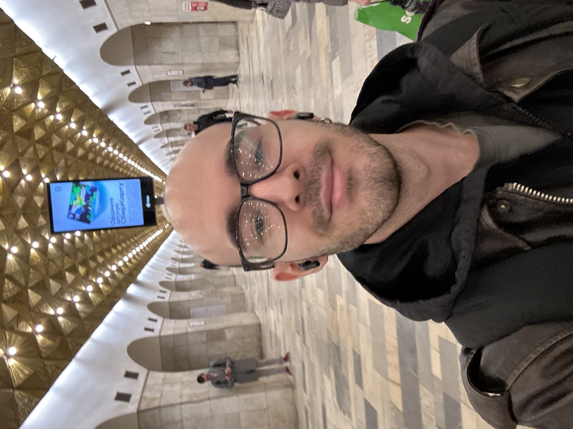 The image is a selfie of a bald man with glasses and a beard. He is wearing a dark jacket and has wireless earbuds in his ears. The man appears to be standing in the Mayakovskaya metro station in Moscow, Russia. The station is known for its Art Deco design, including the vaulted ceilings with geometric patterns and inset lighting. Other people are walking in the background. A blue advertising screen is visible on the left.