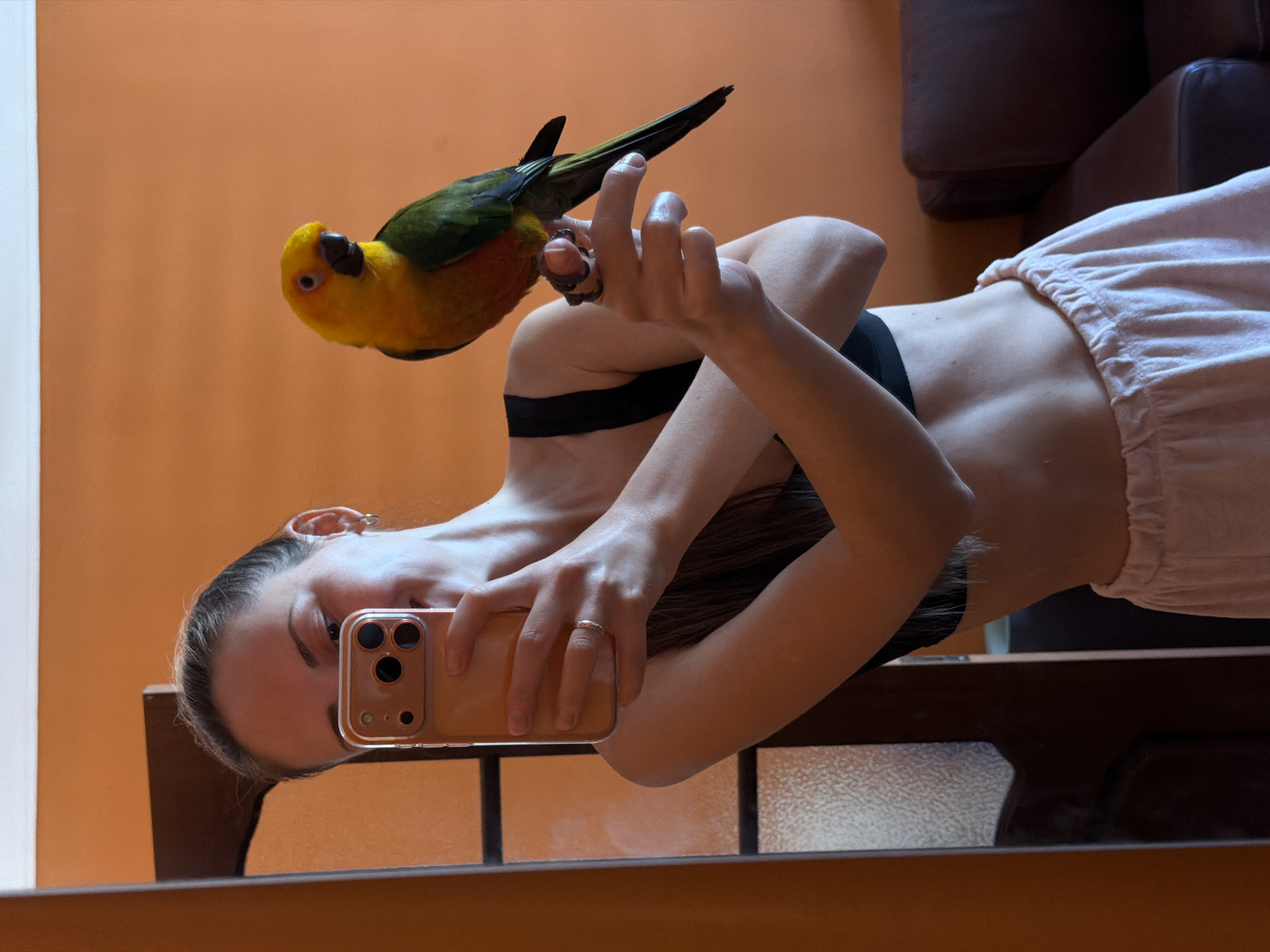 A mirrored image shows a fair-skinned young woman in a black top and light-colored pants holding a smartphone in her right hand, obscuring part of her face. A sun conure parrot sits on her left hand. The background consists of an orange wall and a dark brown leather couch. The image is framed by a mirror border. There is no information available in this image that indicates it was taken in Khimki, Russia.