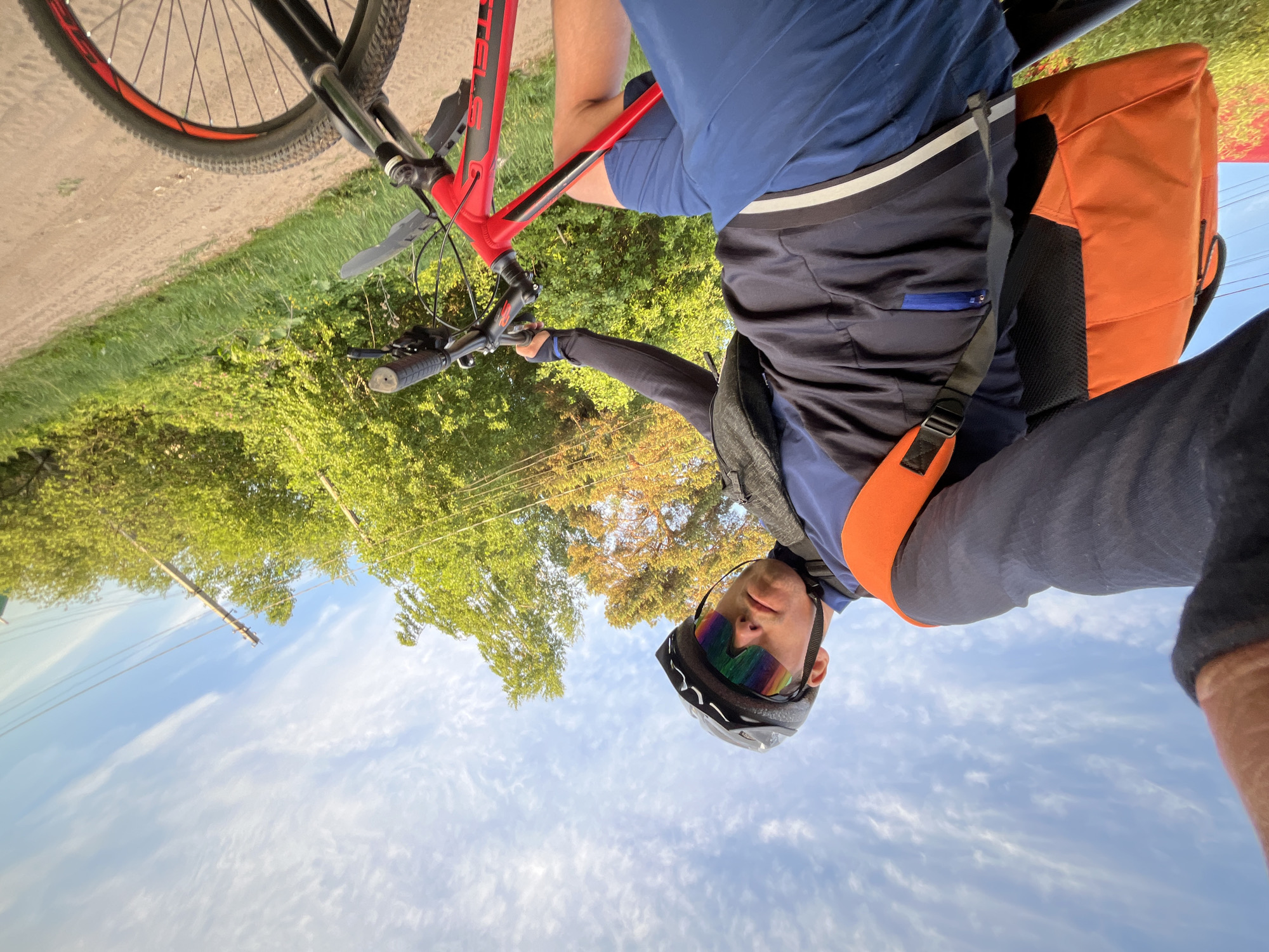 A cyclist is taking a selfie while riding a red and black mountain bike on a road in a grassy area. The cyclist is wearing a blue shirt, black shorts, a helmet, and sunglasses. An orange backpack is visible. The background includes trees and a partly cloudy blue sky. The cyclist is holding the handlebars with one hand and using the other to hold the phone. Power lines can be seen in the background. The location is Chaikovsky, Russia.