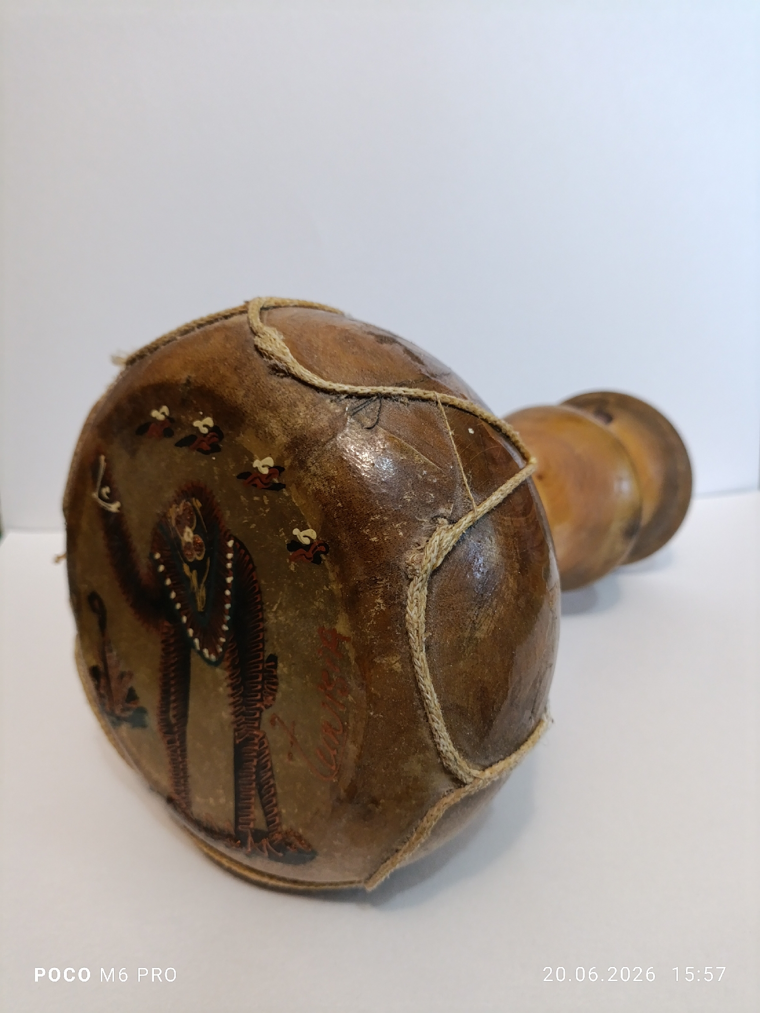 The image displays a handcrafted drum-like object with painted figures on its surface. The object is made of a porous material, possibly animal skin stretched over a gourd, and is bound with twine. The painting depicts two stylized human figures with intricate patterns and what appear to be decorative elements or clothing. Several small, bird-like figures are also painted around the main figures. The object rests on a white surface, likely a table or floor, with a plain white background. The lighting suggests an indoor setting. The timestamp at the bottom right of the image indicates "20.06.2026 15:57", and the bottom left corner displays "POCO M6 PRO". There is no indication of the city of Bender, Moldova, or any specific urban environment within the image itself.