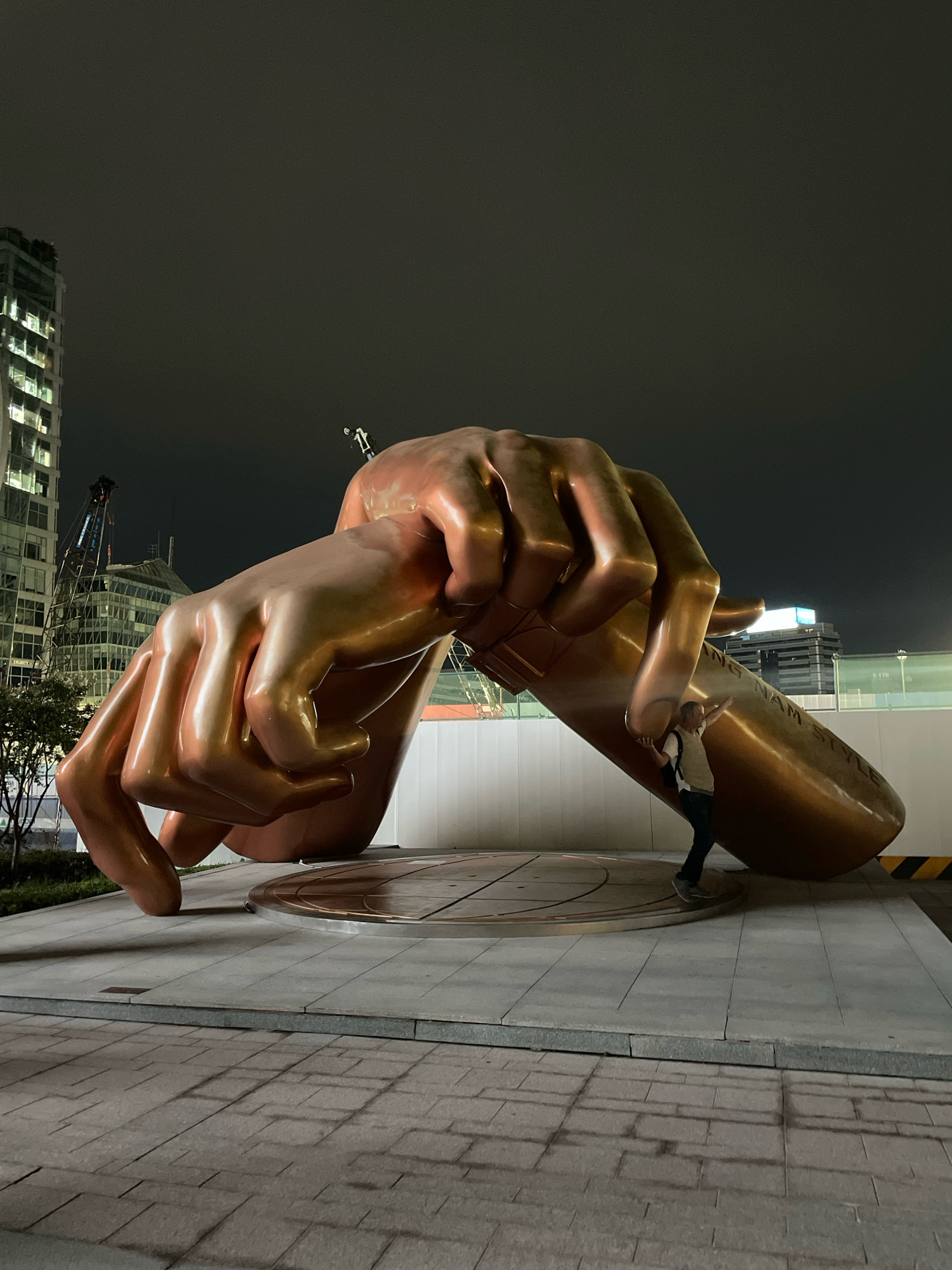 This image depicts a large, bronze sculpture of two hands, seemingly intertwined or grasping, in an outdoor urban setting at night. The sculpture is the focal point, with a person, dressed in a light-colored shirt and dark pants, positioned beneath it, appearing to interact with the base of the sculpture. In the background, modern high-rise buildings are visible, illuminated by interior lights, suggesting an urban environment. Construction cranes and other structures are also present, indicating ongoing development or maintenance in the area. The ground is paved with large, grey tiles and stone blocks. The sky is dark, typical of nighttime. The sculpture appears to be located in the Gangnam district of Seoul, South Korea, near a riverfront area. The inscription "GING NAM STYLE" is visible on the sculpture's base.