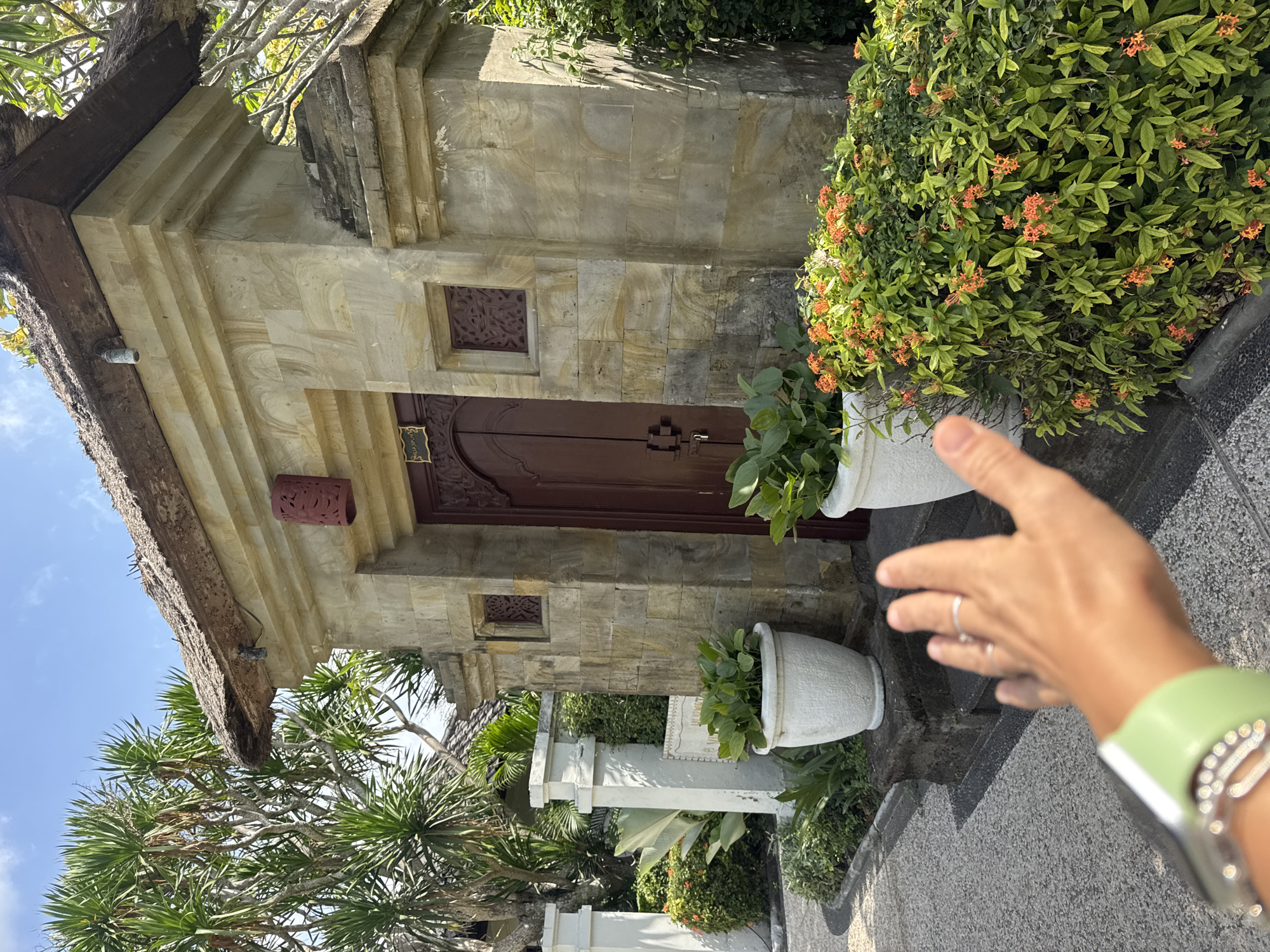 A person's outstretched hand with rings on two fingers and a light green watchband on their wrist is in the foreground of the image, obscuring part of the view of a stone building. The building has a thatched roof, ornate wooden doors painted dark red, and decorative carved stone insets. Two large white planters with green foliage sit on stone steps leading to the entrance. Lush green vegetation with small orange flowers is to the right of the building. To the left, a paved walkway leads past white pillars and more greenery. The setting appears to be outdoors in a tropical location, likely a resort or temple complex in Kuta Selatan, Indonesia, given the architectural style and surrounding foliage. The sky is blue with some clouds.