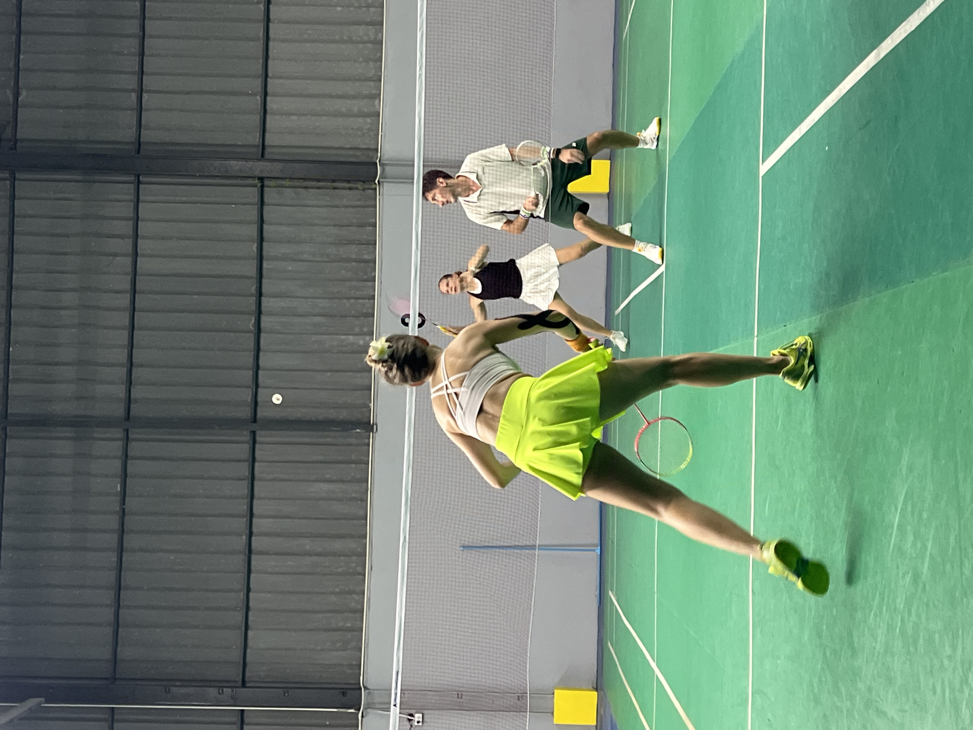 Four individuals are playing badminton on an indoor court with a green surface and white lines. A net divides the court. In the foreground, a woman in a white sports bra and a bright yellow skirt is in motion, facing away from the camera and holding a badminton racket. Behind her, a woman in a black tank top and white skirt is positioned to hit the shuttlecock. To her right, a man in a plaid shirt and green shorts is also holding a racket, looking towards the shuttlecock. Further back, a fourth person, mostly obscured by the net and other players, is also present. The background consists of a dark, corrugated metal wall. The lighting appears to be artificial. The court is marked with yellow rectangular objects on either side of the net, and blue posts support the net. This scene is taking place at a badminton court, likely in the vicinity of Ban Rang Rak, Thailand, suggested by the context of the prompt.