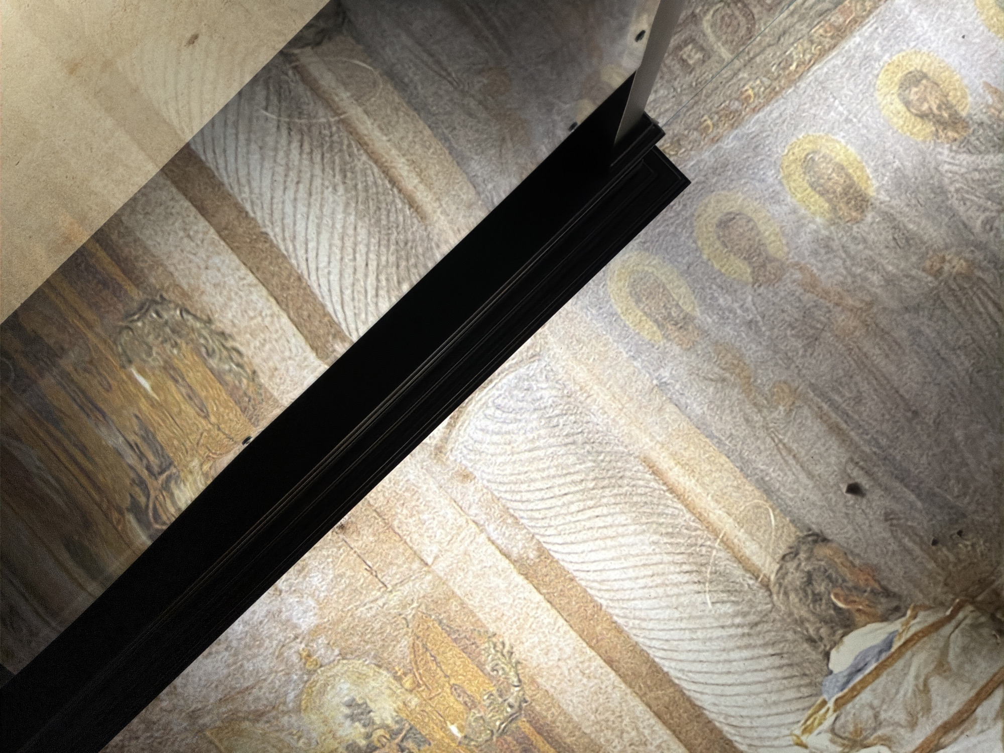 A low-angle view shows a fragment of an ornate ceiling, likely part of a historical or religious building. The ceiling is adorned with frescoes or murals depicting figures with halos, suggesting religious iconography. The artwork appears faded or aged. A thick, black, rectangular frame or beam is diagonally bisecting the image, providing a strong contrast to the intricate detail of the ceiling. A metallic rod appears to be supporting the frame. The location is in Sofia, Bulgaria.