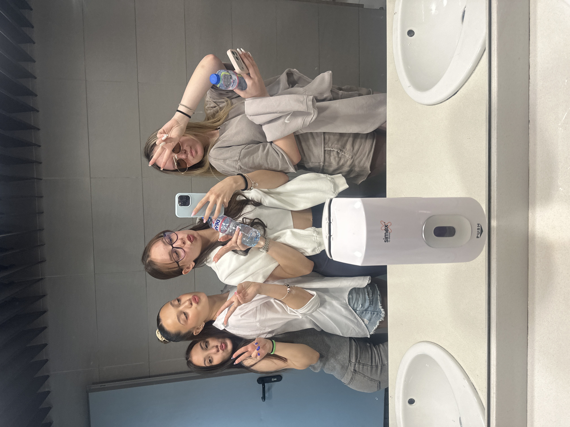 Three females are taking a mirror selfie in a public restroom. The mirror reflects the three subjects and the top portion of a fourth individual. The subject on the left wears glasses and is holding a water bottle while the subject in the center is wearing a white blouse and holding her fingers in a peace sign. The subject to the right is wearing sunglasses and holding her fingers in a peace sign, and is holding a water bottle. A soap dispenser and two sinks are also present. The location is undetermined.
