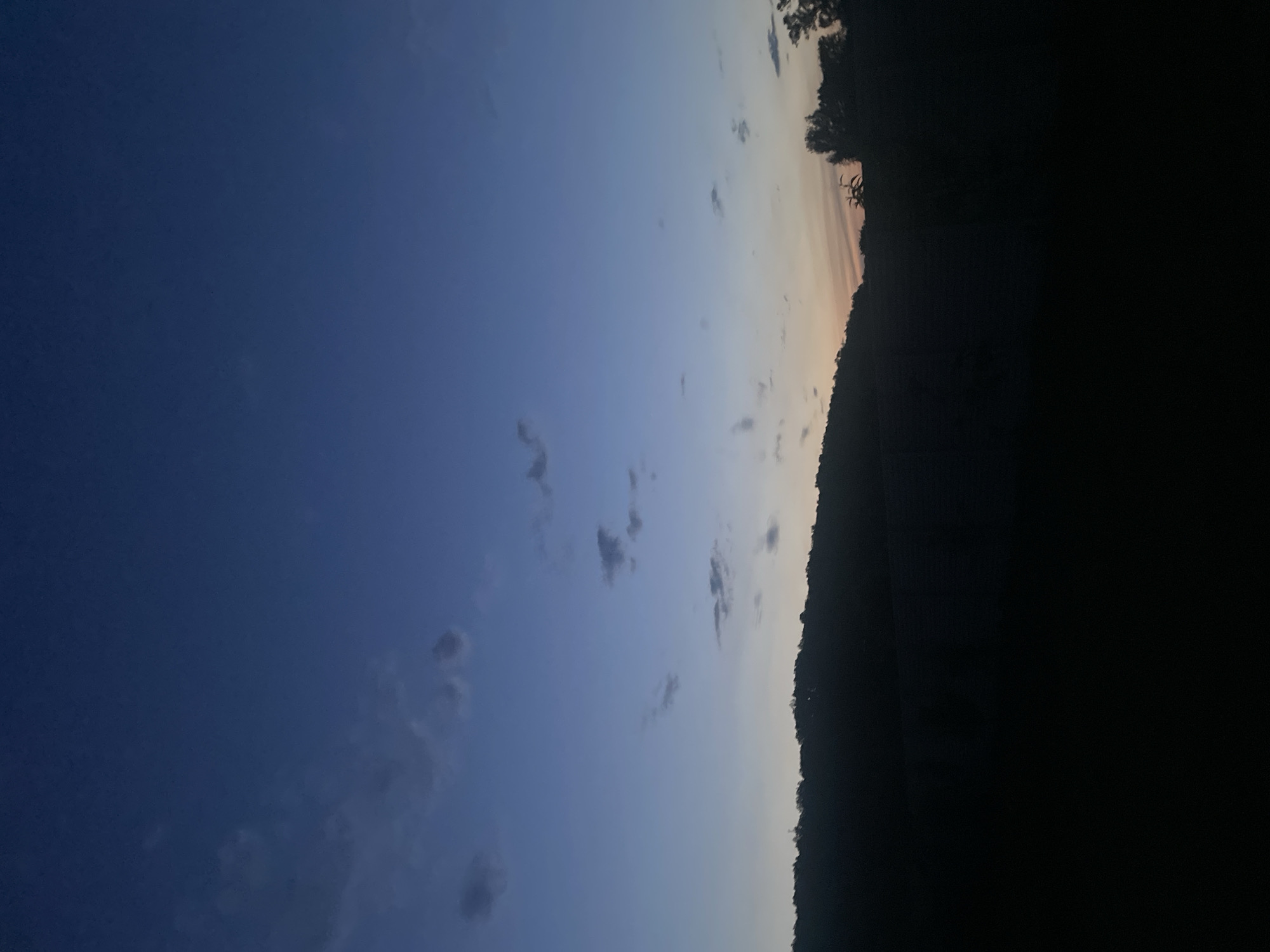 The scene depicts an outdoor landscape, observed during twilight or dawn, in the presumed location of Petrivka, Ukraine. The sky occupies approximately the upper two-thirds of the vertical frame, displaying a color gradient that transitions from a dark blue at the zenith to a lighter blue in the middle sky, culminating in a narrow band of faint orange and yellow light along the horizon. Several small, dark gray clouds are scattered across the blue portions of the sky, primarily in the upper left and central areas. A dark, undulating silhouette, consistent with a distant treeline or hilly terrain, forms the horizon line. The lower one-third of the image is uniformly dark, obscuring any specific details of foreground features or ground cover. No human figures, animals, man-made objects, or specific actions or interactions are discernible within the frame.