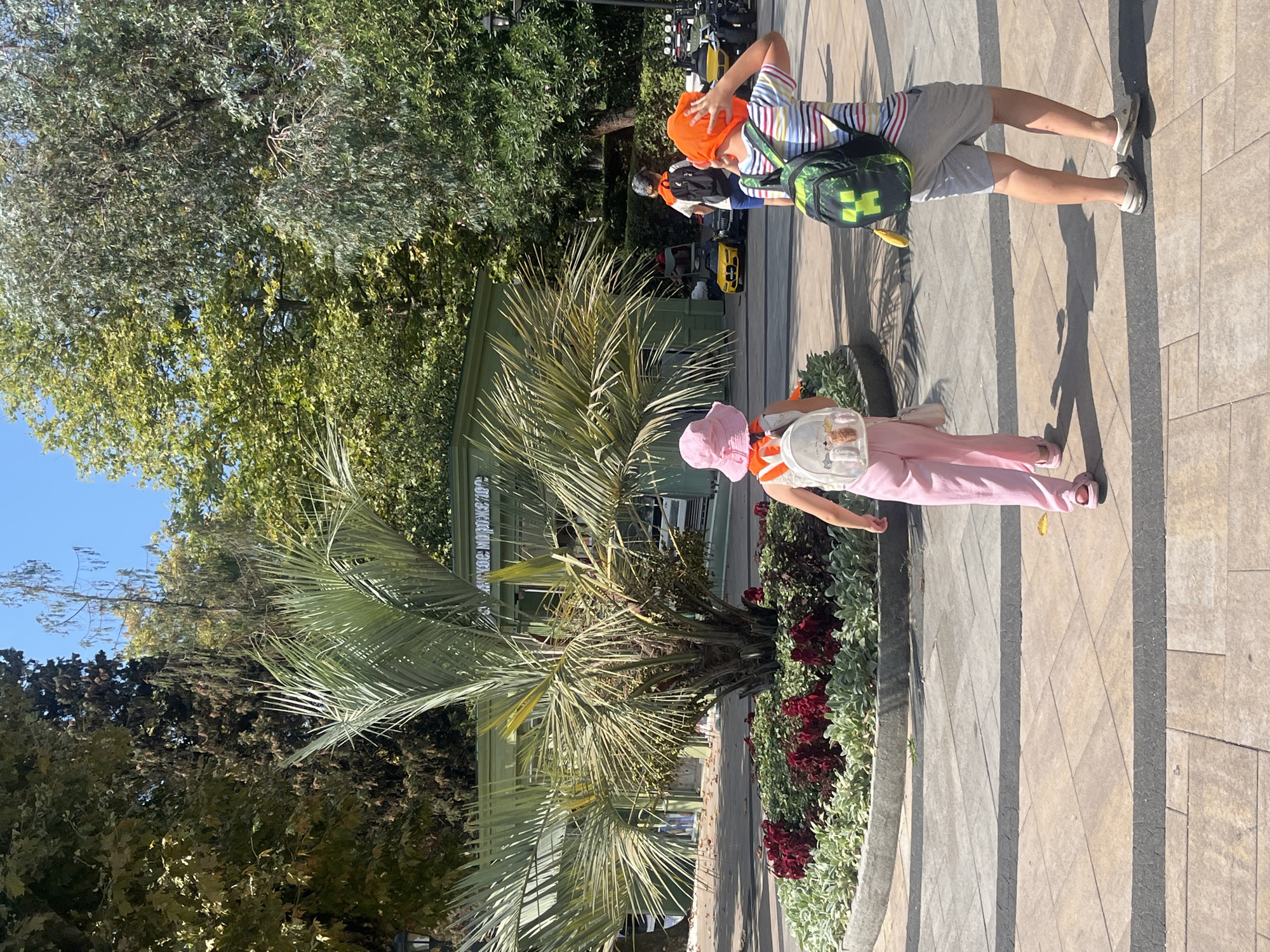 In this medium shot, two children are walking in a park on a sunny day. The child on the left is a girl wearing a pink bucket hat and pink pants, carrying a white backpack with cartoon characters. The child on the right is a boy wearing an orange beanie and a striped t-shirt with gray shorts, carrying a green backpack with a yellow Minecraft character. They are walking on a paved path with geometric patterns. In the background, there is a palm tree, flower beds with red and green foliage, and a green building with Cyrillic text. The location is likely a public park or promenade in Sochi, Russia, given the palm tree and the Cyrillic text on the building. The overall atmosphere is casual and recreational, suggesting a family outing or a visit to a tourist attraction.