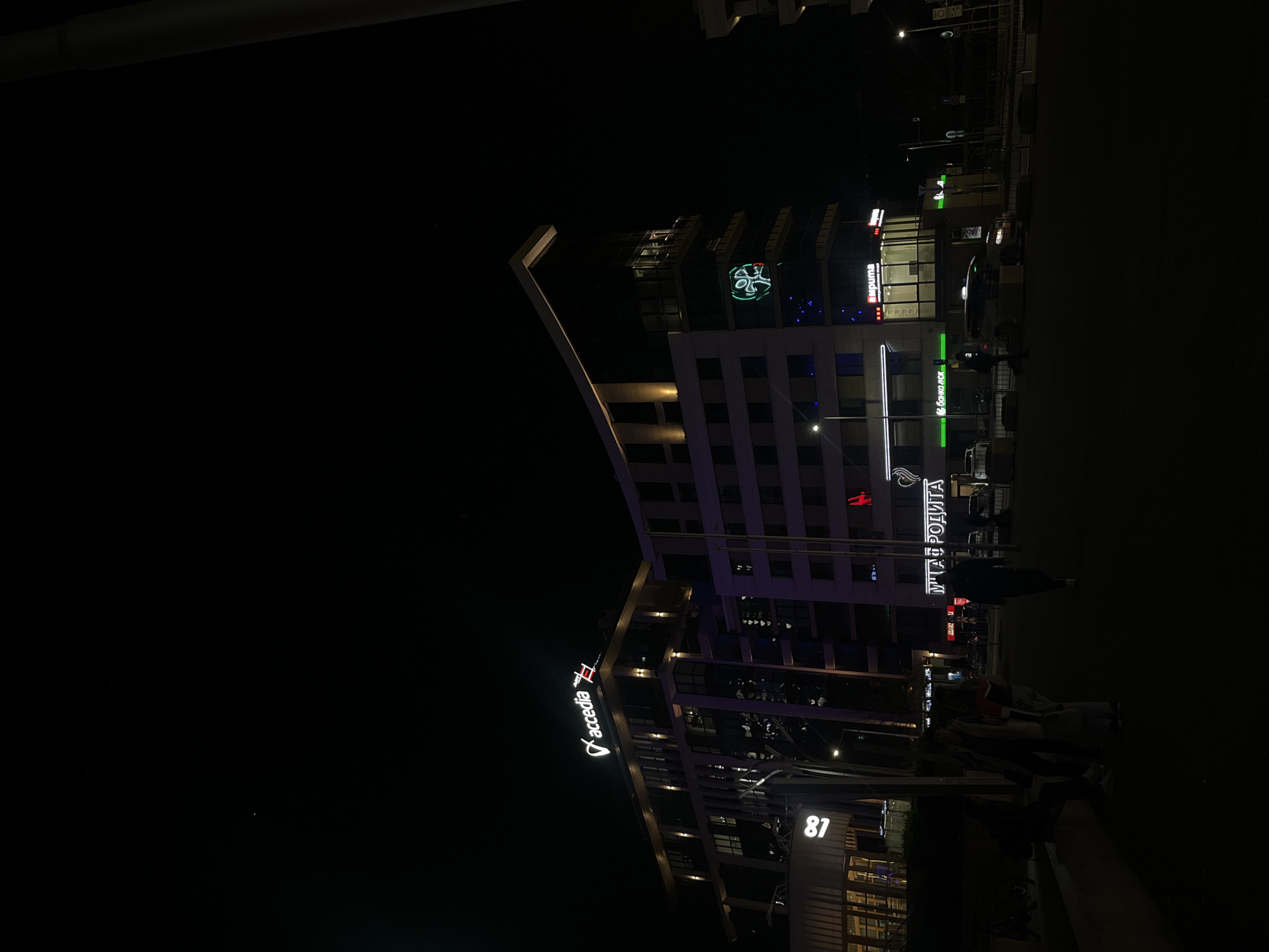 The image shows a nighttime view of a building in Sofia, Bulgaria. The building has several visible business signs, including "Accedia," "МЛАДОСТА," and a cloverleaf logo. People are present on the sidewalk in front of the building. The address "81" is visible on the building's facade. Other illuminated signs and streetlights are also present.