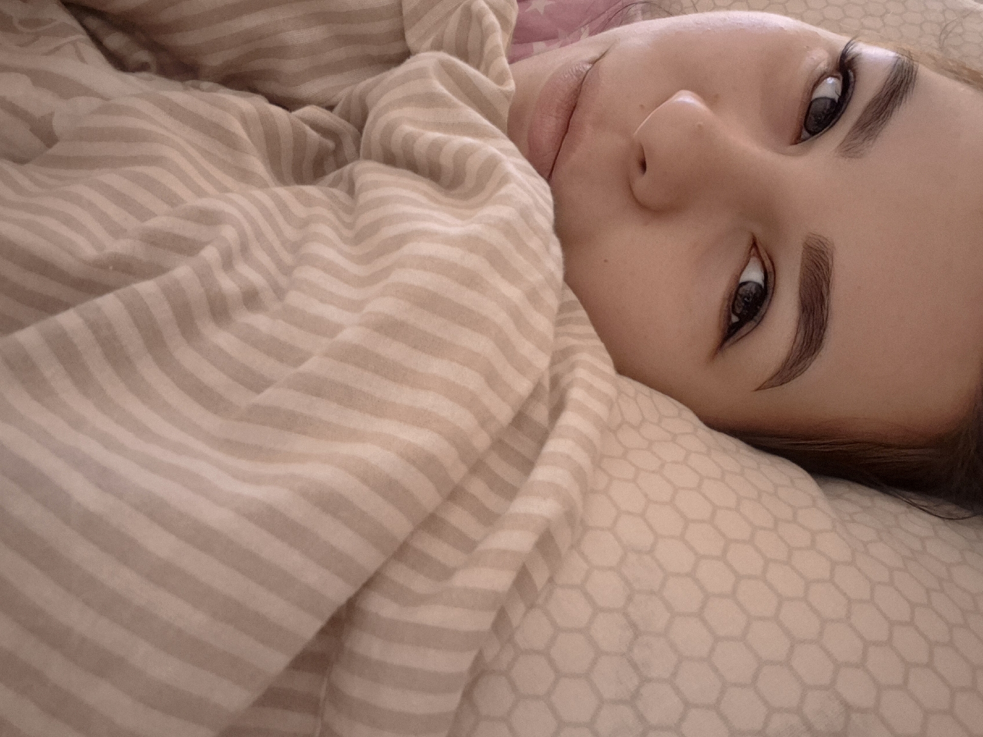 A young woman is lying in bed, partially covered by a striped blanket. Her face is in focus, showing her eyes, nose, and mouth. She appears to be resting on a pillow with a hexagonal pattern. The environment suggests a domestic setting. There is no evidence to suggest that this image was taken in Tobolsk, Russia.
