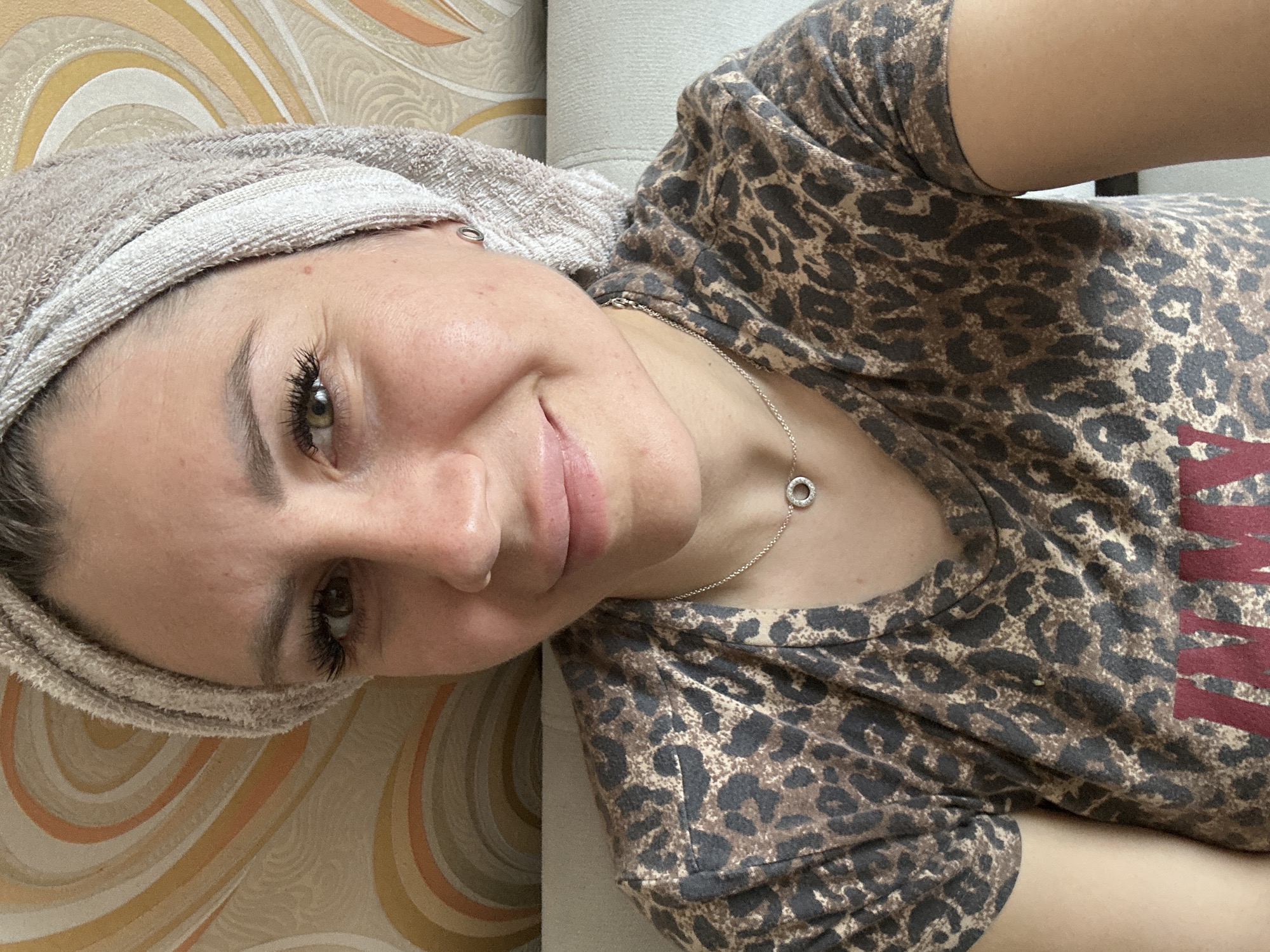 A woman is shown from the chest up, wearing a leopard-print t-shirt and a beige towel wrapped around her head. She is smiling and looking towards the camera. She is wearing a delicate silver necklace with a circular pendant. The background appears to be a wall with a patterned wallpaper. The lighting suggests an indoor setting. There is no information available to determine the specific location within the city of Novoaltaysk, Russia.