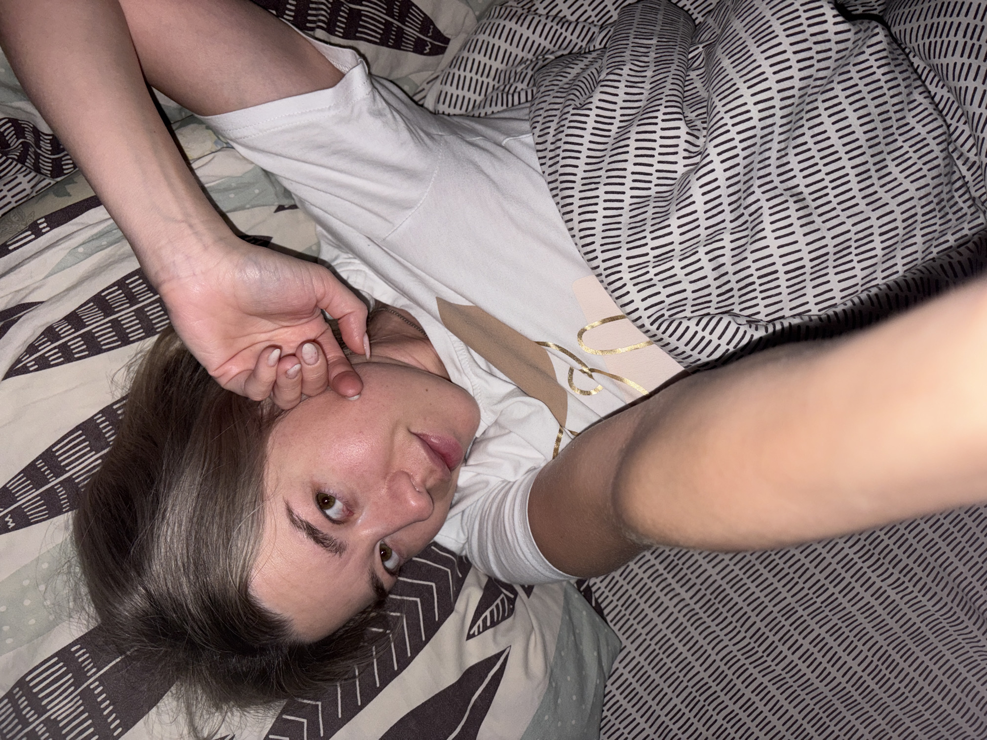 A young woman with shoulder-length light brown hair lies on a bed with patterned sheets and a patterned comforter. She is wearing a white t-shirt with a design on the front. Her left arm is raised and her hand is near her face. Her right arm is extended towards the camera, creating a selfie-like perspective. The location is unknown, but the query includes "city of Chaikovsky, Russia" as a potential location.