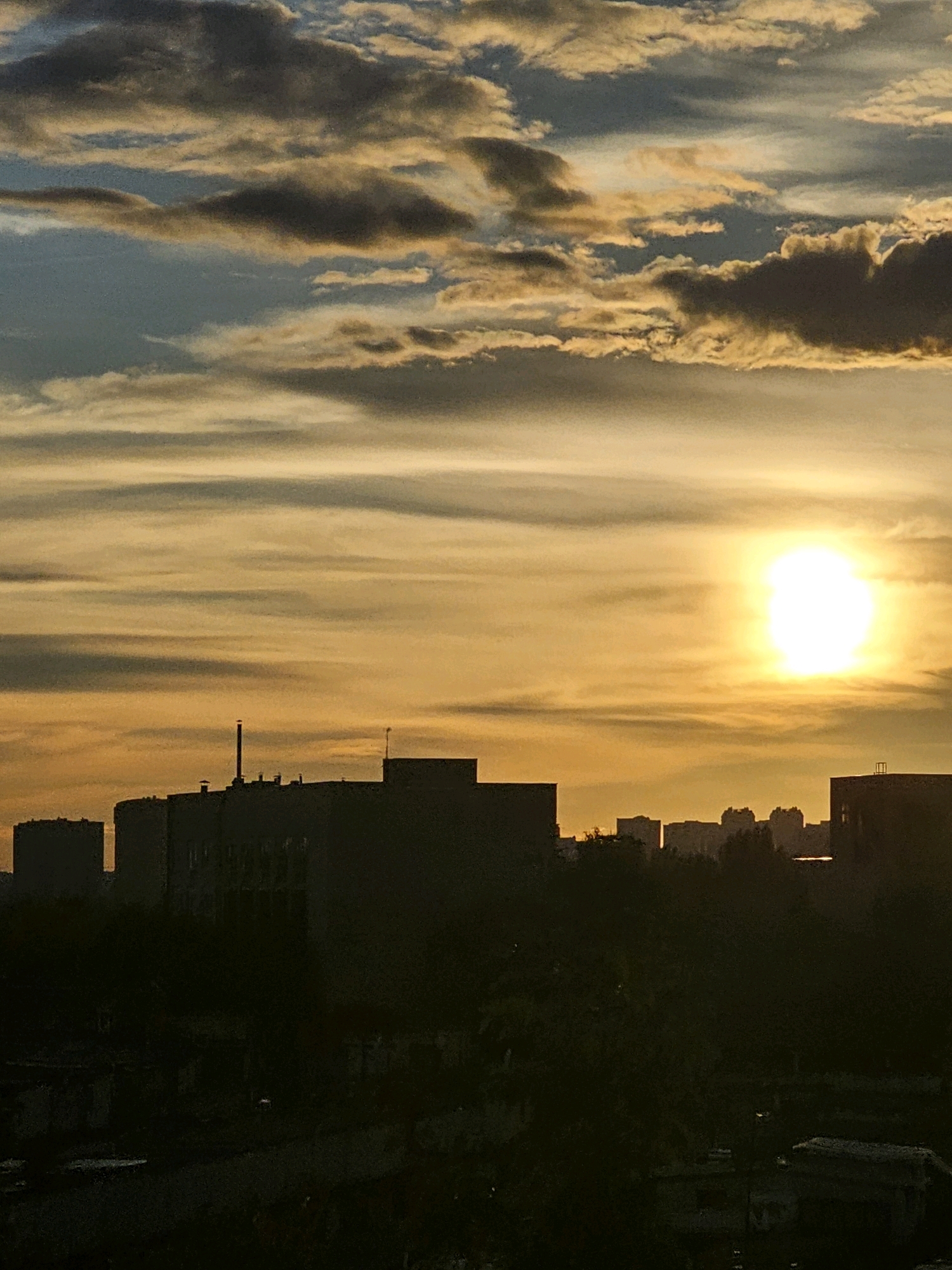 This is a landscape photograph taken at sunset in the city of Khimki, Russia. The foreground is dominated by the dark silhouettes of buildings and trees against the warm glow of the setting sun. The sky is filled with a mix of soft, wispy clouds colored in shades of orange, yellow, and blue. The sun, a bright white orb, is prominently positioned in the upper right quadrant of the frame, casting its light across the clouds and illuminating the city below. The overall mood of the image is serene and atmospheric, capturing the beauty of a sunset over an urban environment.