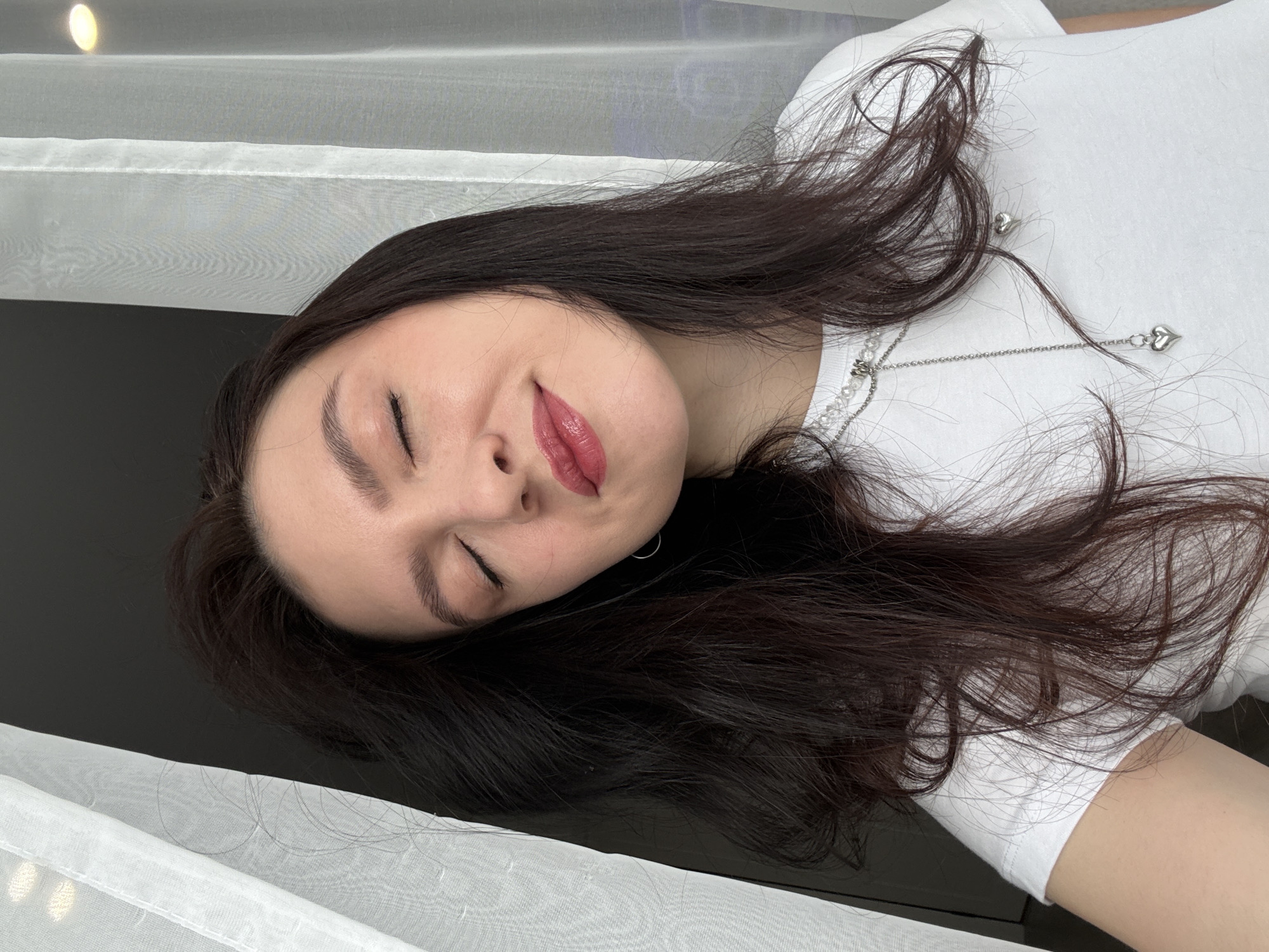 A woman with dark hair lies on a dark, glossy surface next to a white curtain. She is wearing a white t-shirt and a silver necklace with a heart pendant. Her eyes are closed and she has a slight smile. The location in the city of Kazan, Russia is not discernible from the image.