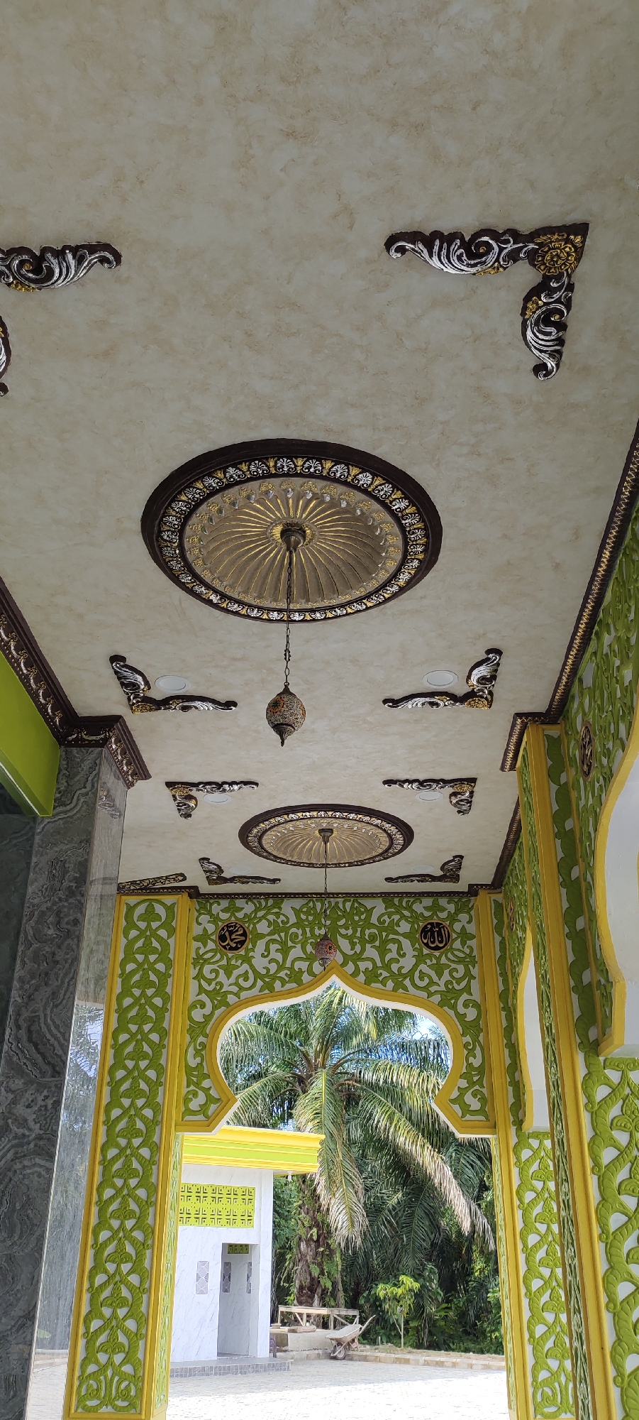 The image shows the interior ceiling and arched doorway of a building in Aluekeujruen, Indonesia. The ceiling is decorated with ornate circular motifs and corner accents featuring a mix of brown, gold, and silver. Two pendant lamps hang from the ceiling. The arched doorway, framed by vibrant green and yellow decorative patterns, opens to an outdoor scene with palm trees and lush vegetation. A small building with white walls and green decorative elements is visible through the archway.