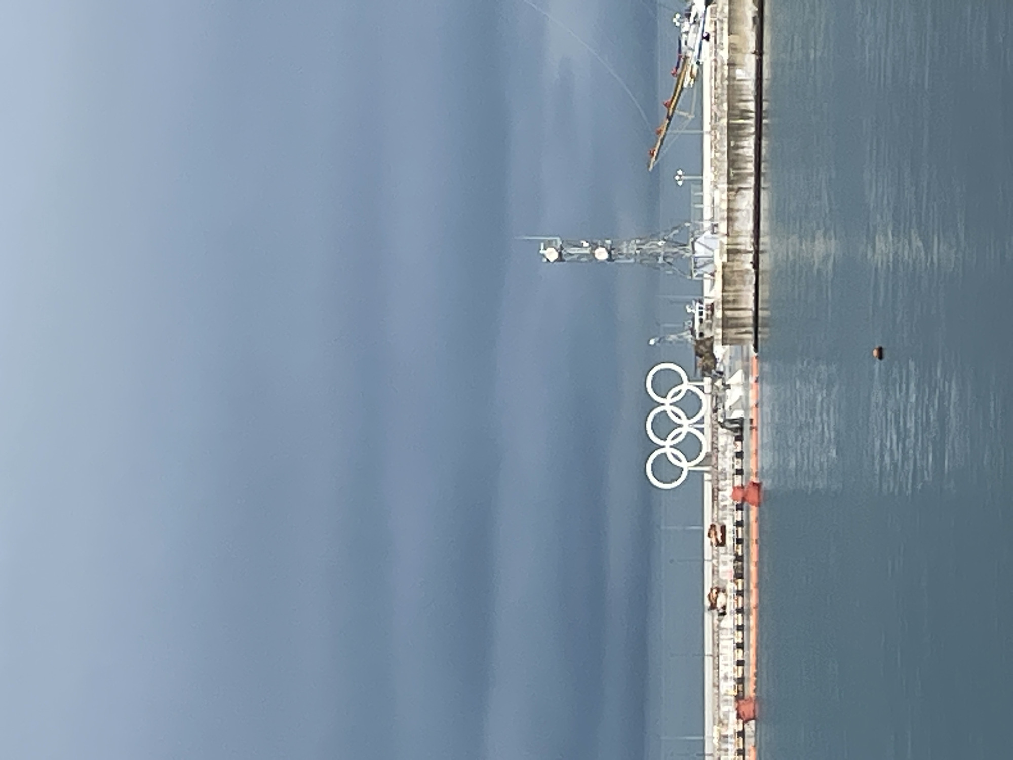 This image shows the Olympic rings at the port of Sochi, Russia, next to the water. The rings are large and white, set against a cloudy sky. A pier or dock stretches along the water's edge with various structures and equipment visible, including a tower. An orange buoy floats in the water. There are small figures of people visible walking along the dock. The sky is overcast.