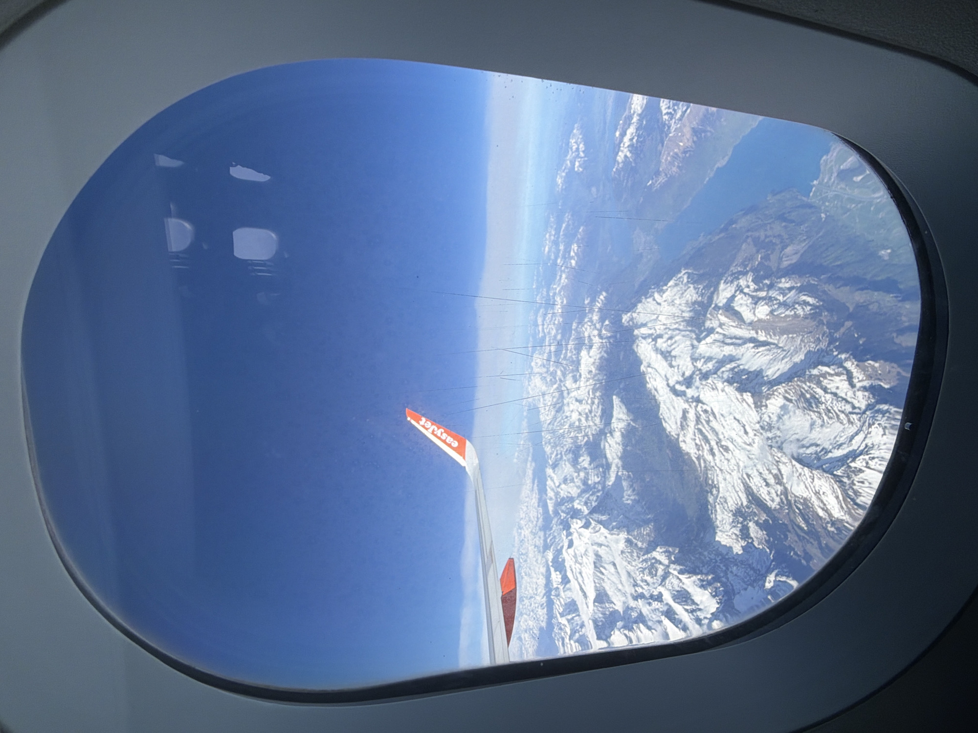 The image presents a high-altitude view from an airplane window, showcasing a vast natural landscape. The primary subjects are the airplane's interior window frame, a portion of the aircraft wing, and the expansive scenery outside.
The view is framed by a grey, oval airplane window, which exhibits noticeable scratches on its surface and some internal reflections, particularly in the upper left area, appearing as bright white shapes. A prominent feature is the airplane's wing, visible in the lower center and extending upwards towards the middle. The wing has a light grey surface, and its upturned winglet is a striking orange with the word "easyJet" clearly printed in white text.
Beyond the wing, the scene reveals a clear, deep blue sky dominating the left side of the frame, indicating bright, sunny daytime conditions with no clouds visible. The right and central portions of the view are filled with a magnificent range of snow-capped mountains. These rugged peaks and valleys are covered in white, suggesting a high-altitude flight over an alpine region. To the upper right, a large body of deep blue water, likely a lake or a significant waterway, is visible, bordered by mountainous terrain.
The image captures an in-flight moment, likely over the European Alps, given the provided context of Milan, Italy, showing the beauty of mountainous landscapes from above.