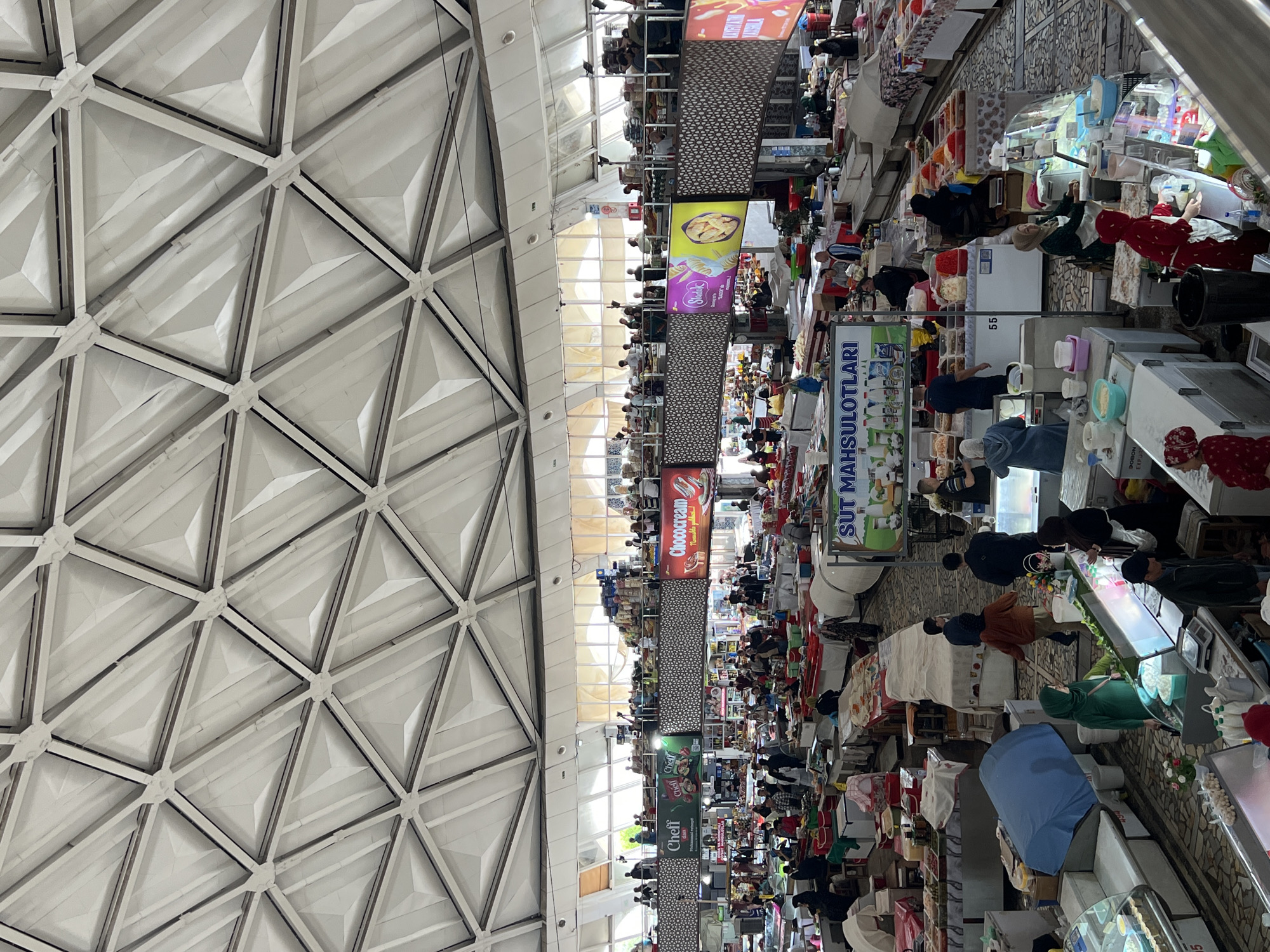 This indoor shot shows the bustling atmosphere of the Chorsu Bazaar in Tashkent, Uzbekistan. The perspective is unusual, presenting the scene sideways with the ceiling on the left. The ceiling has a geometric, triangular pattern, while the main section features numerous stalls arranged in rows, full of merchandise and crowded with people. Individuals, dressed in a variety of colors, can be seen browsing and interacting with the vendors. There are various goods on display, including what seems to be food, fabrics, and other wares, with banners and signs hanging above the stalls. The scene conveys a vibrant sense of commerce and cultural exchange within a large indoor market.