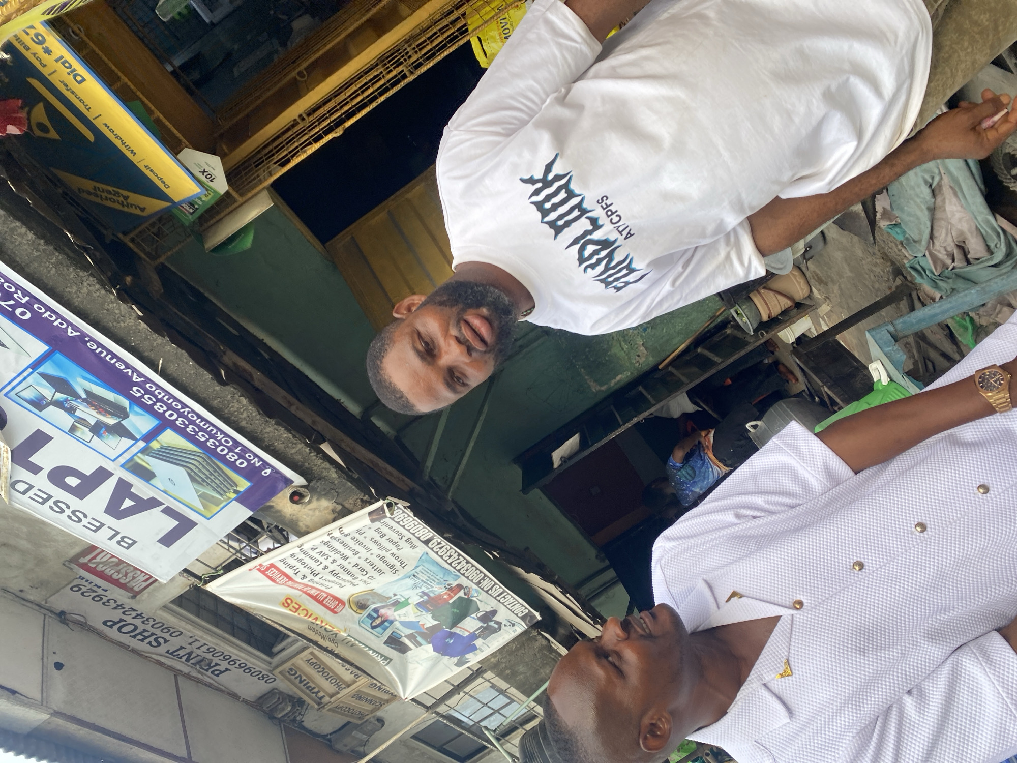 Two men stand together in Lagos, Nigeria, under a canopy of shop signs that advertise services such as printing, photocopying, passport photos, and typing. One man wears a white t-shirt with the word "FUNDLOOK" printed on it, while the other is dressed in a white textured jacket with gold buttons. The environment includes signs advertising businesses like "BLESSED LAPT" and "PRINT SHOP," and indicates phone numbers for contacting these services. The "Dial *67" sign suggests mobile banking transactions are also facilitated here.