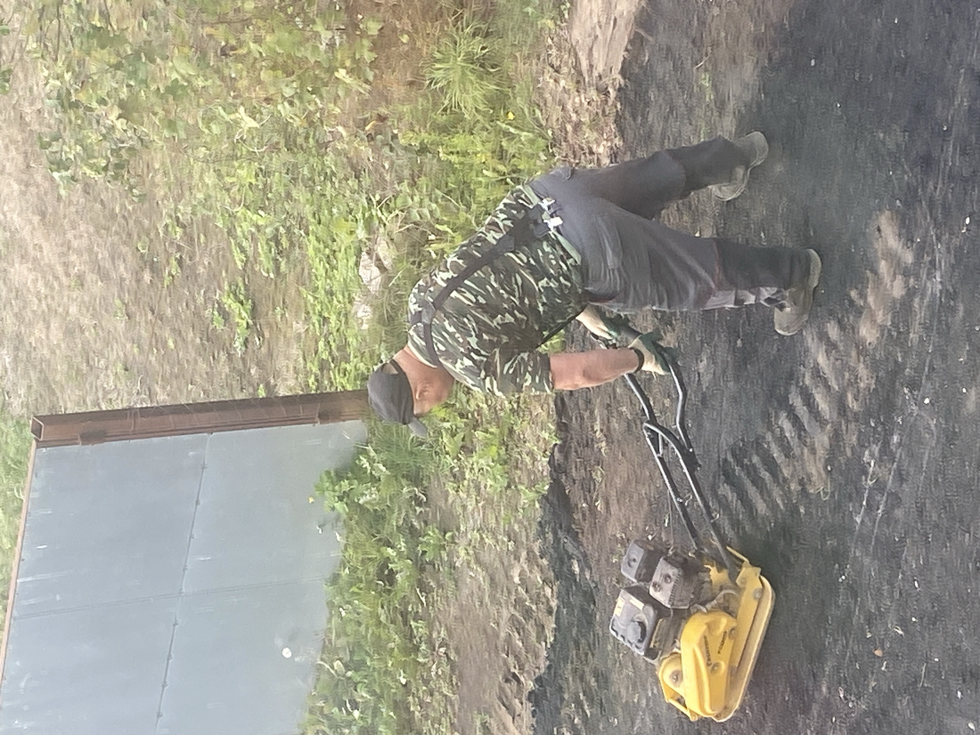 In Krupki, Belarus, a person wearing a camouflage shirt, gray pants, a black baseball cap, and work boots is operating a yellow and black vibratory plate compactor. The compactor is positioned on a dark, loose material that has been recently worked. The individual is holding the handles of the compactor, guiding it as it compacts the surface, leaving visible parallel indentations. In the background, a large, light gray metal fence or wall stands vertically, supported by a rusty metal post. Beyond the fence, a patch of green vegetation and dry, uneven ground are visible. The overall scene suggests ongoing construction or landscaping work.