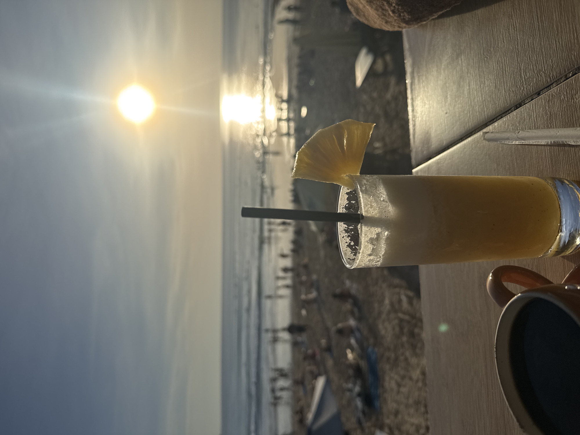 The image captures a scene on a beach, likely in Kuta Selatan, Indonesia, during sunset. The sun is setting over the ocean, creating a bright, reflective glare on the water. A crowded beach is visible in the background. In the foreground, a glass of yellow smoothie, garnished with a slice of pineapple and a straw, sits on a wooden table. A mug, partially visible, is positioned next to the glass.
