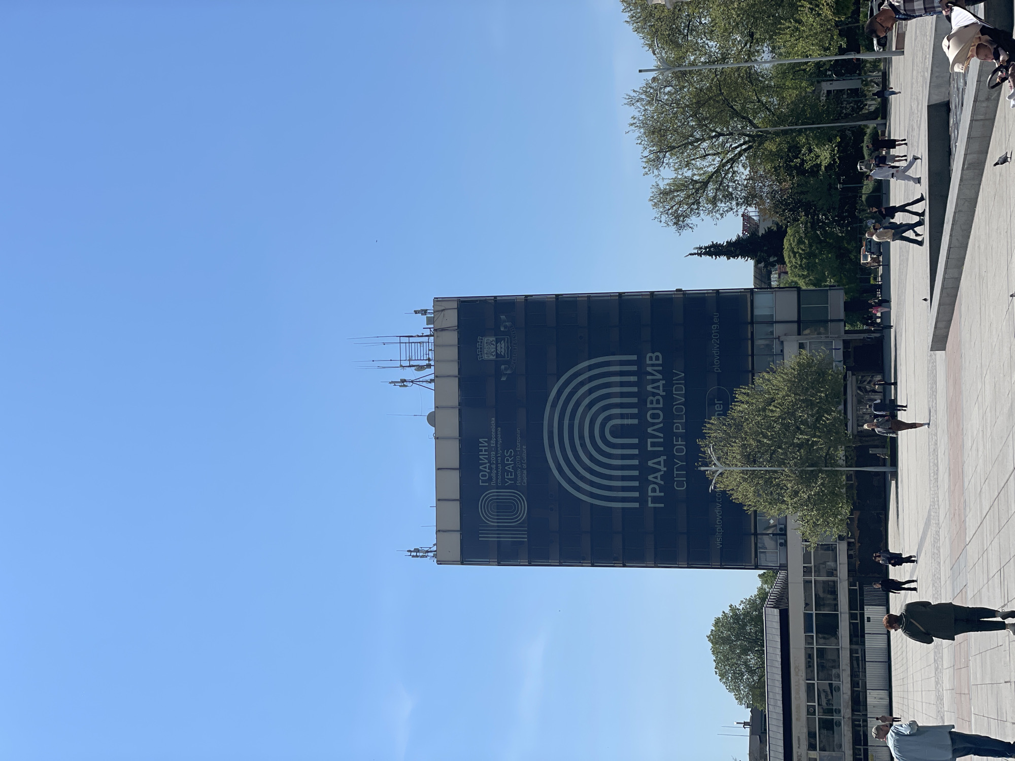The image depicts an outdoor urban scene in Plovdiv, Bulgaria, under a clear, bright blue sky. A large, modern building with a dark glass facade dominates the left side of the frame, featuring a prominent banner or screen. This banner displays text in Bulgarian and English, reading "ГОДИНИ" (YEARS) and "Plovdiv 2019 - European Capital of Culture," alongside a large "10" graphic, suggesting an anniversary or commemoration. Further down, "ГРАД ПЛОВДИВ" (CITY OF PLOVDIV) is shown with a rainbow-like arched logo, and the websites "visitplovdiv.com" and "plovdiv2019.eu" are visible. The top of the building is fitted with various antennas and communication structures.
In the foreground and to the right, a spacious, paved public square or plaza is filled with people. Various individuals and small groups are seen walking or standing, dressed in casual clothing. Their distinct shadows on the ground indicate strong, direct sunlight, suggesting it is daytime, likely morning or early afternoon. Lush green trees are interspersed throughout the plaza and in the background, providing shade and adding a natural element to the urban environment. Several other buildings of varying sizes are visible behind the trees, contributing to the cityscape. The overall impression is that of a lively and active public space in Plovdiv.