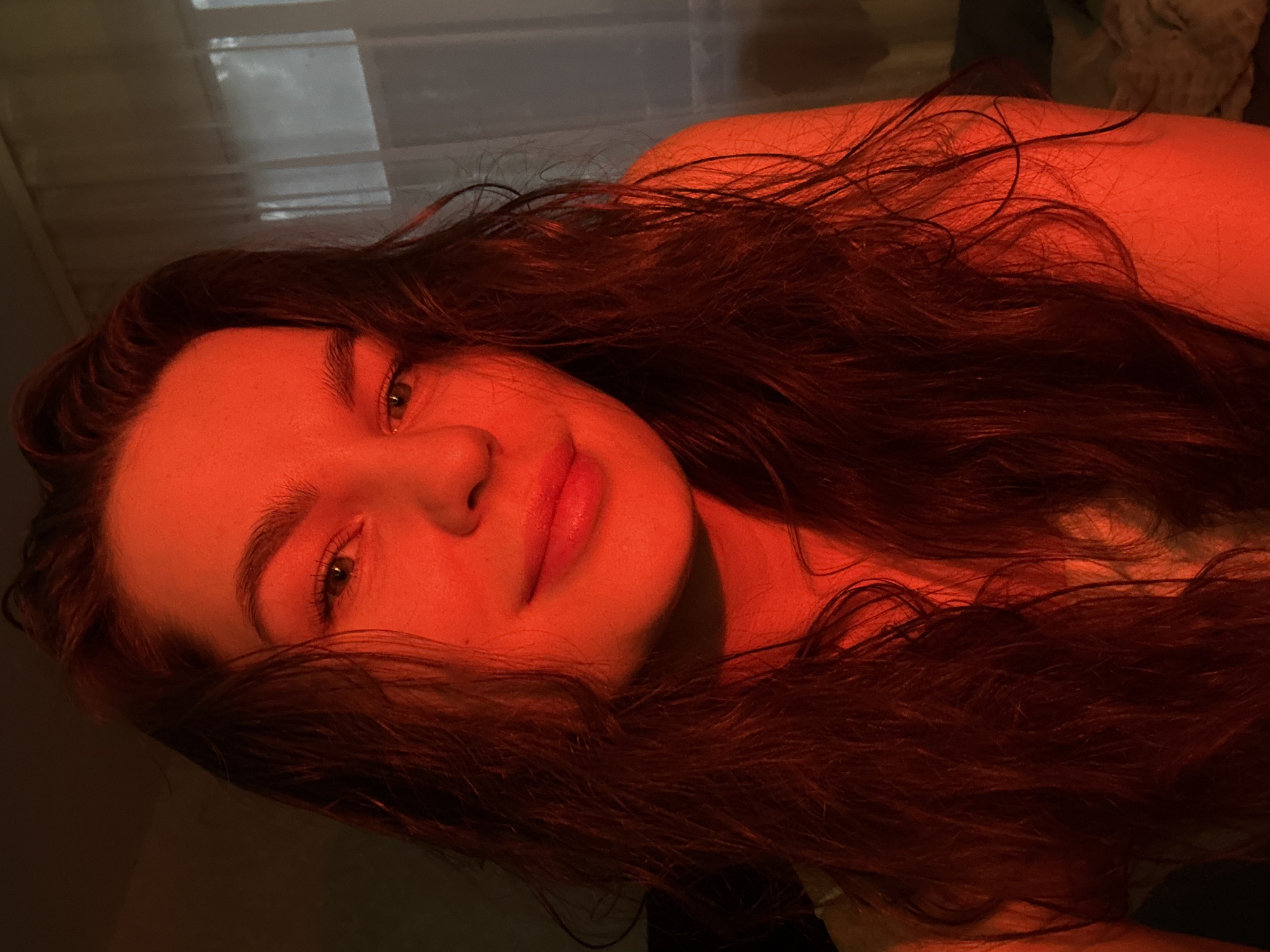 The image is a close-up, head-and-shoulders selfie of a young woman with long, dark, wavy hair. She has fair skin and is illuminated by a strong red light source, casting a warm, dramatic glow across her face and hair. Her eyes are open and looking towards the camera, and she has a subtle smile. Her eyebrows are dark and well-defined, and she appears to be wearing makeup on her lips.
The background is blurred, but suggests an indoor setting with a window covered by sheer curtains on the right side. The lighting suggests the sun may be setting, or an artificial red light is being used.
This image was taken in Chaikovsky, Russia.